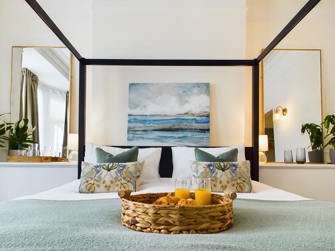 Bright modern bedroom with four-poster bed, teal accents, wall art, mirrors, and wicker tray on the duvet