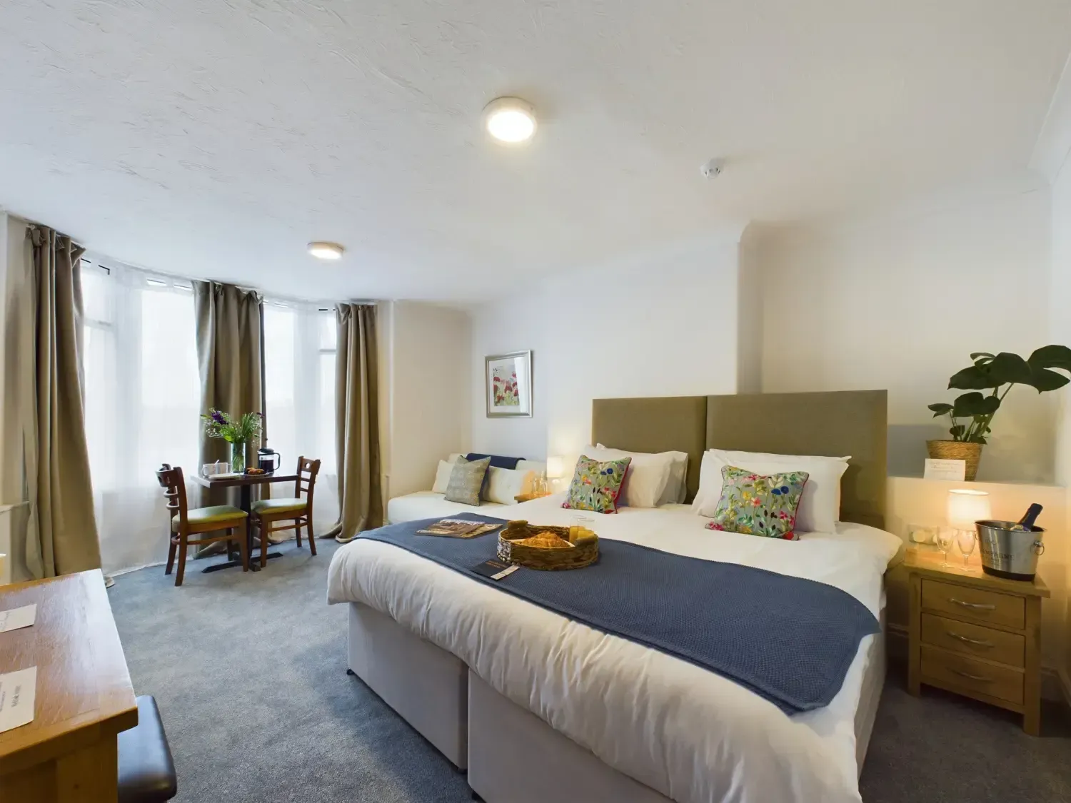 Bright bedroom with a large bed, white walls, gray carpet, and a small dining table by the windows.