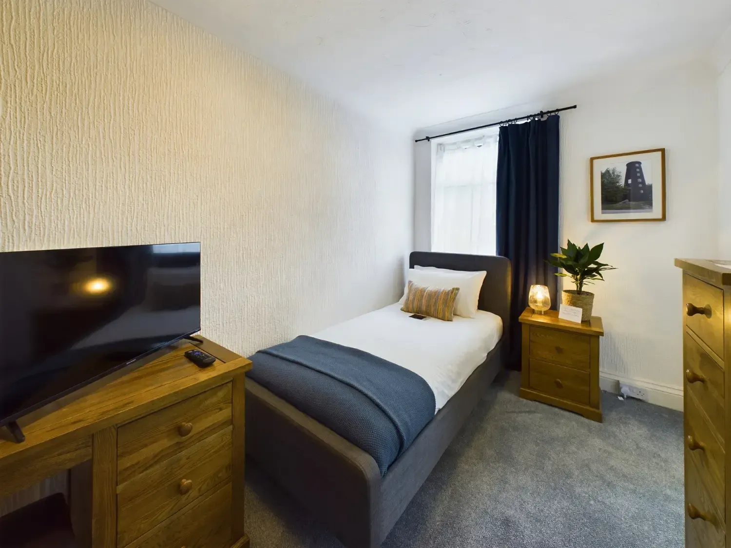 Small bedroom with a single bed, wooden furniture, TV, and window with dark curtains.