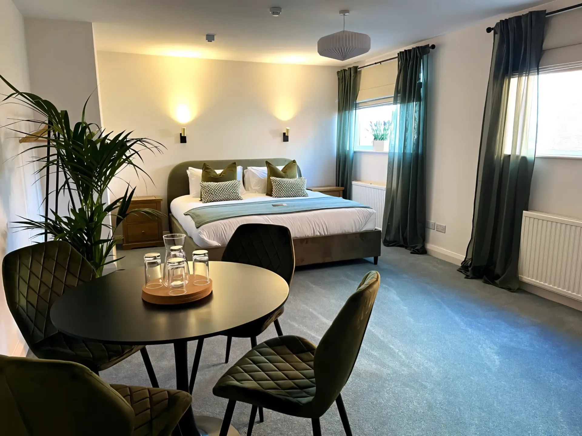 Cozy hotel bedroom with twin beds, round table, plants, and large windows with curtains
