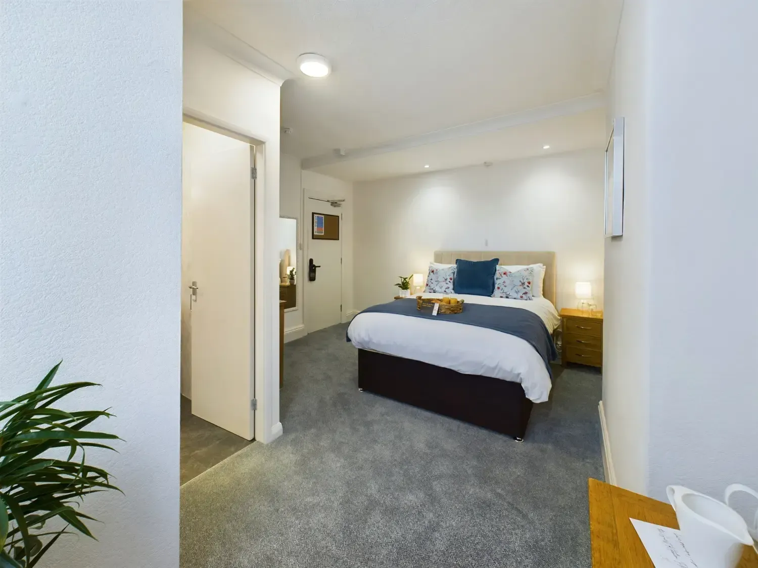 Bright bedroom with a neatly made bed, gray carpet, and warm bedside lamps.