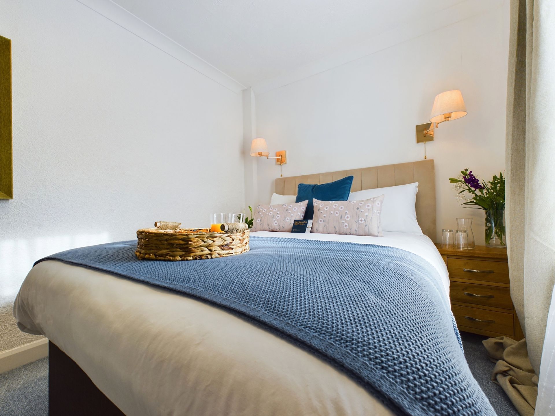 Bright bedroom with a neatly made bed, blue knit throw, bedside lamps, and a small wooden nightstand.
