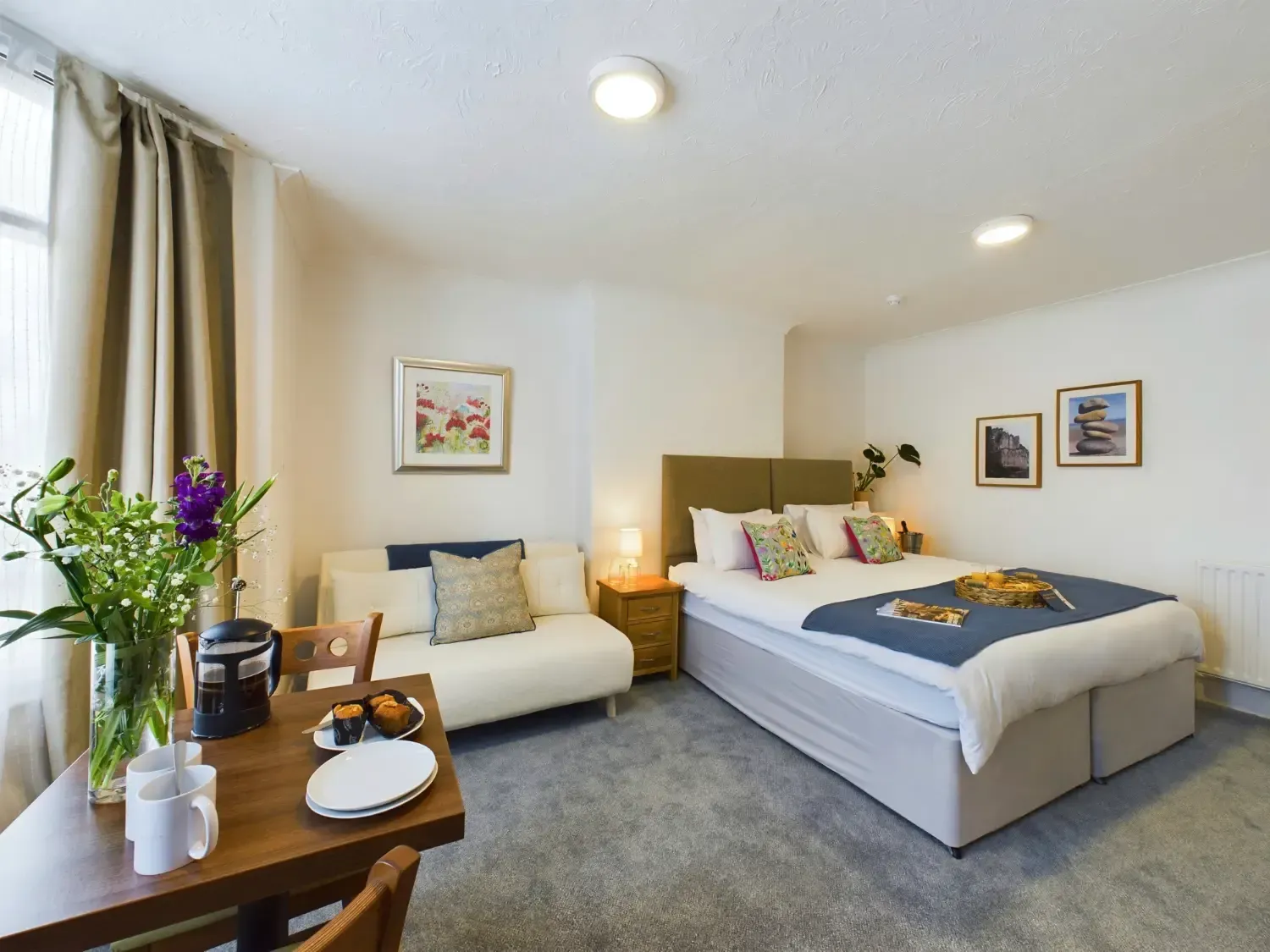 Bright bedroom with a neatly made bed, armchair, desk, lamp, and wall art in soft neutral tones.