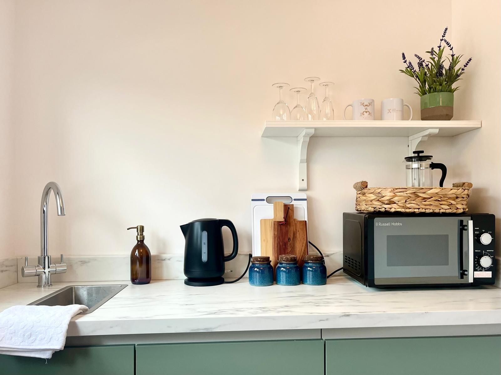 White kitchen counter with sink, kettle, microwave, jars, and a small potted plant on a shelf.