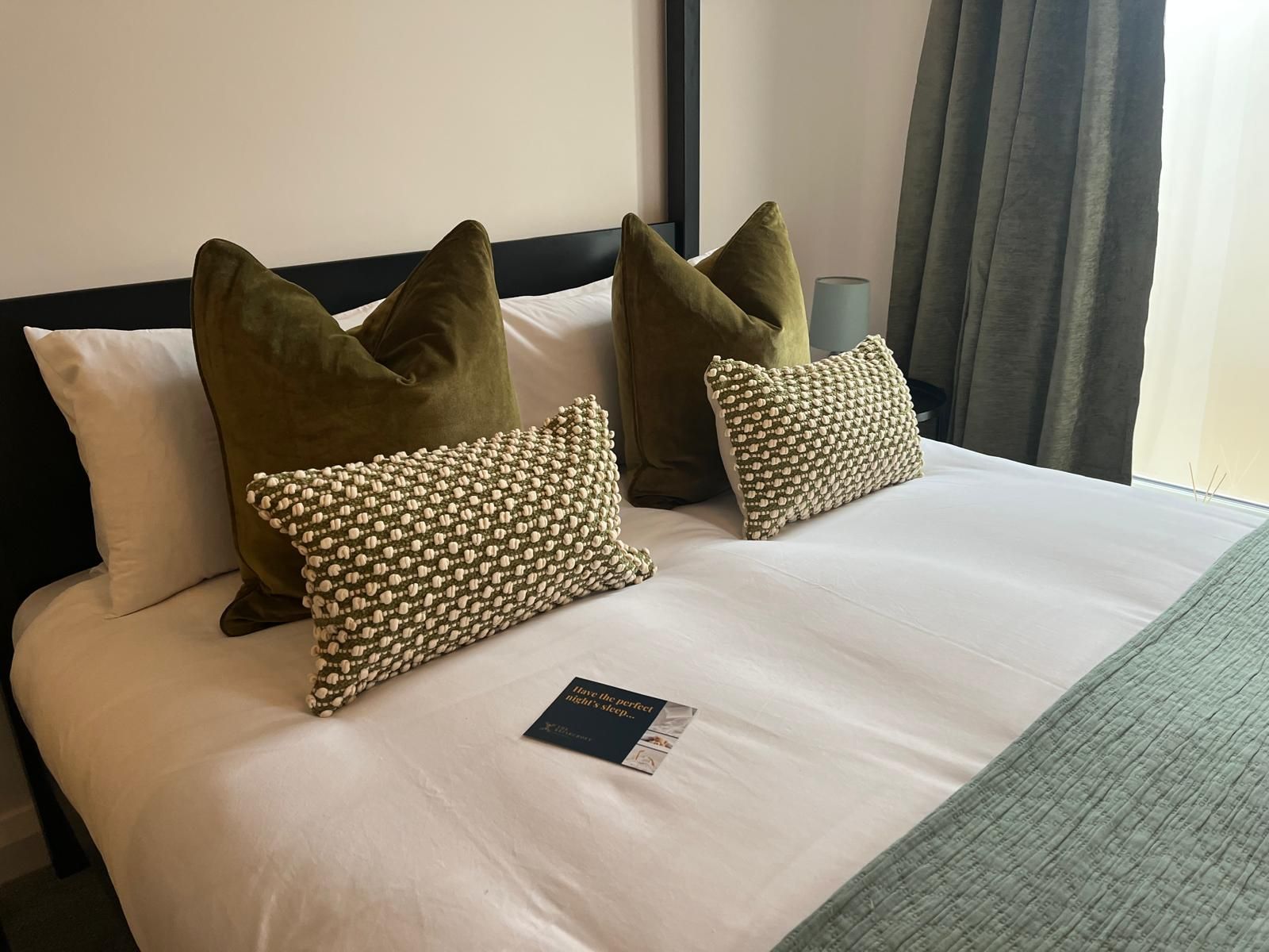 Neatly made bed with white bedding, olive and patterned pillows, and a green throw near a window