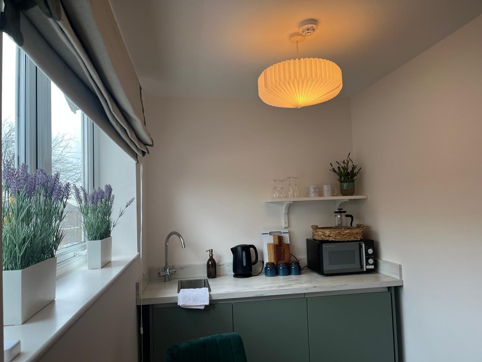 Modern kitchen corner with green cabinets, white countertop, and window herb planters under a pendant light