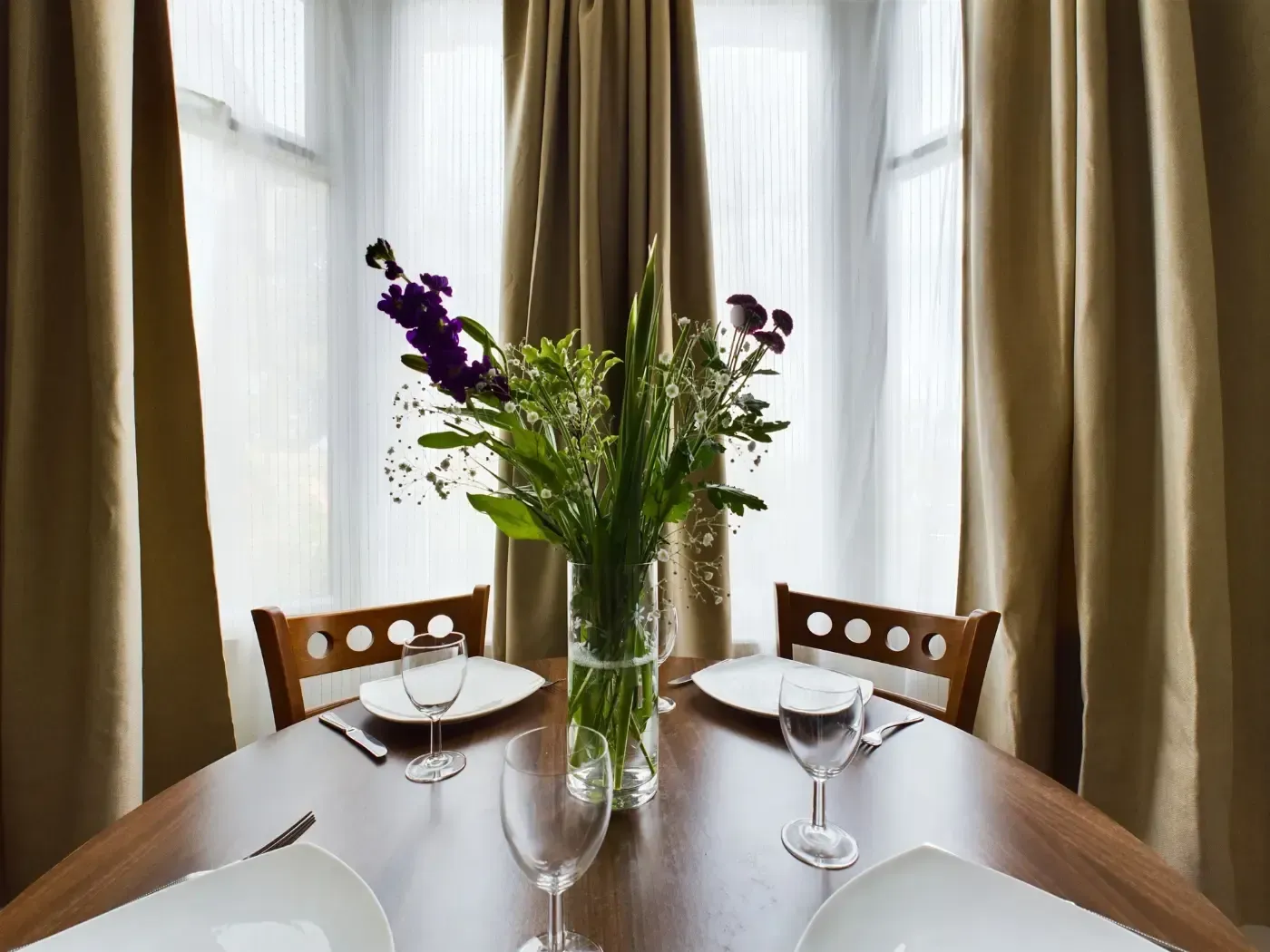 Dining table set for two by bright windows with curtains and a vase of flowers in the center