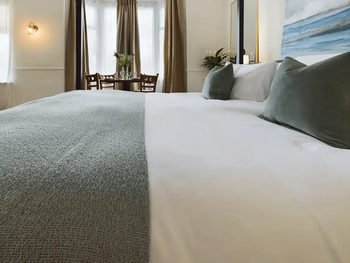 Neatly made bed with gray blanket and pillows in a bright bedroom near a window and small desk