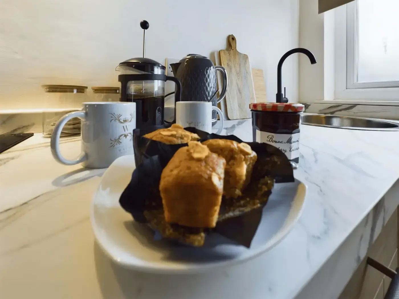 Plate of muffins on a kitchen counter with a mug, coffee maker, and window in the background