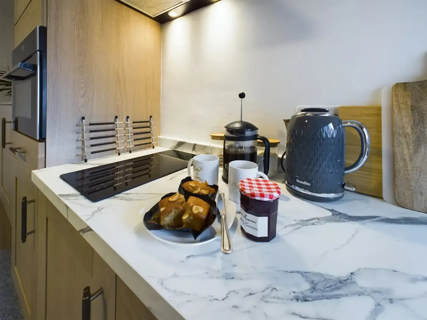 Modern kitchen countertop with stovetop, blue kettle, French press, bread, and jam jar