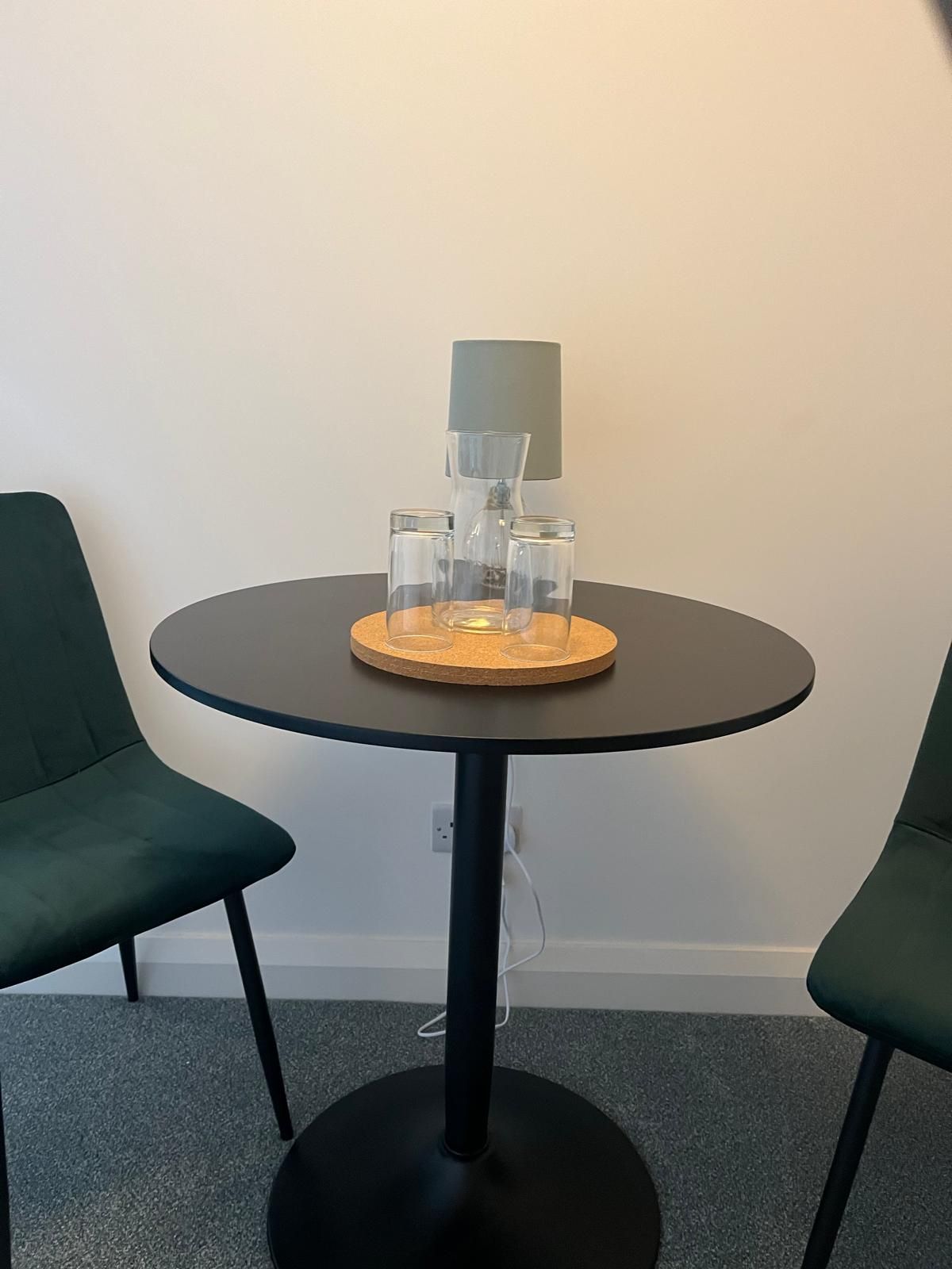 Small round table with two green chairs, glasses on a wooden tray in a minimal room.