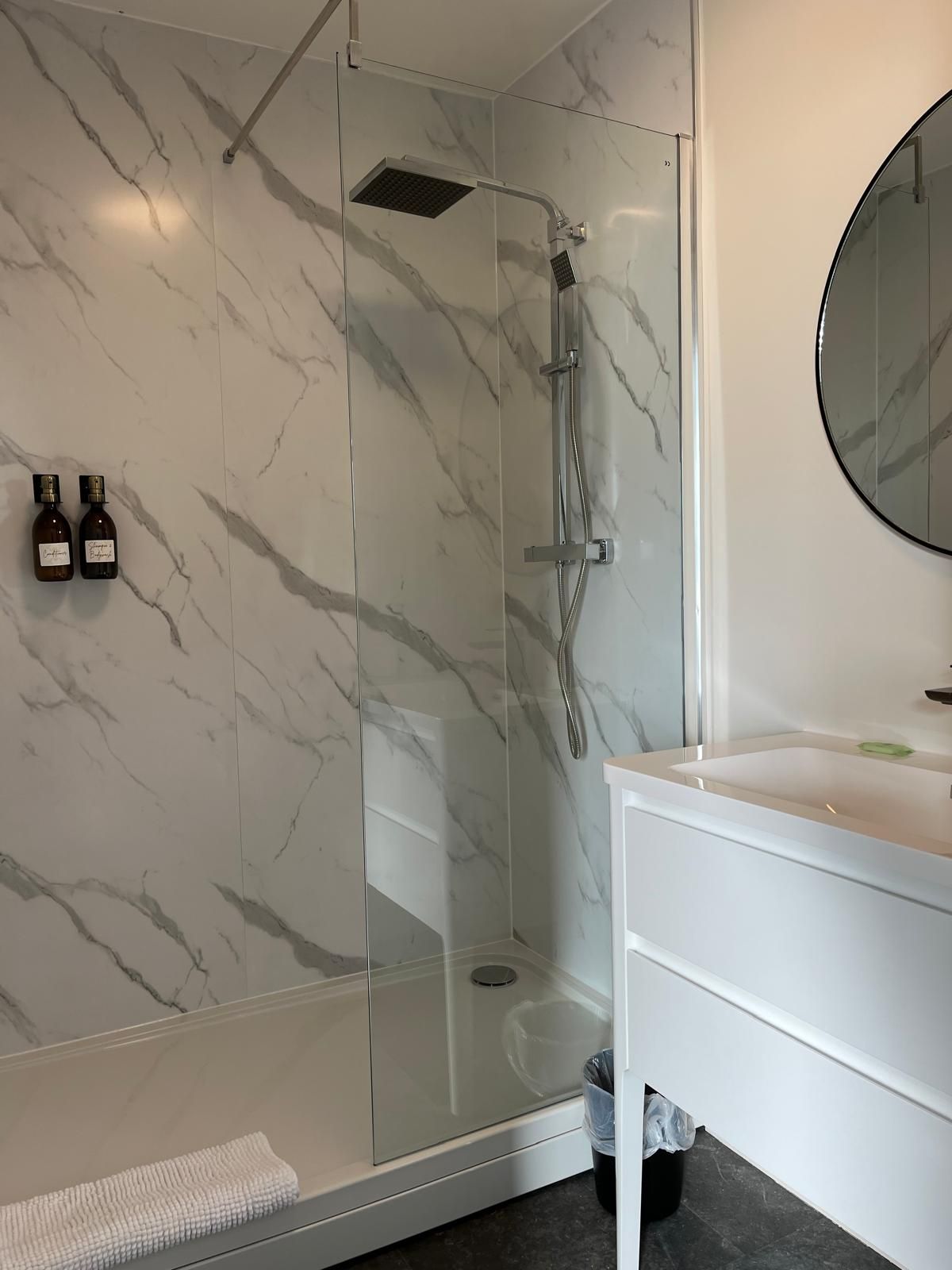 Modern bathroom with marble shower, glass door, white vanity, and round mirror