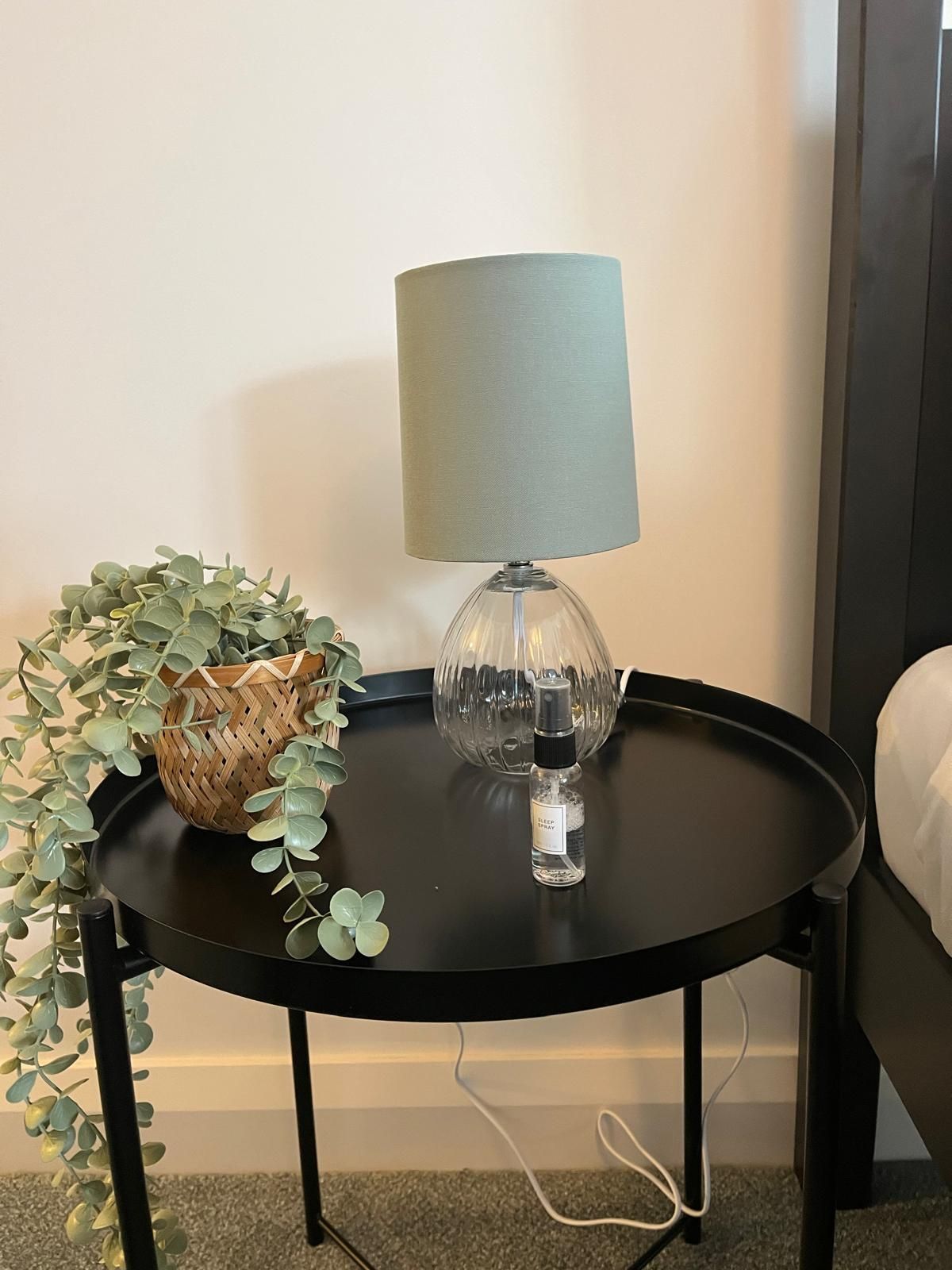 Black round side table with lamp, plant, and glass decor against a beige wall