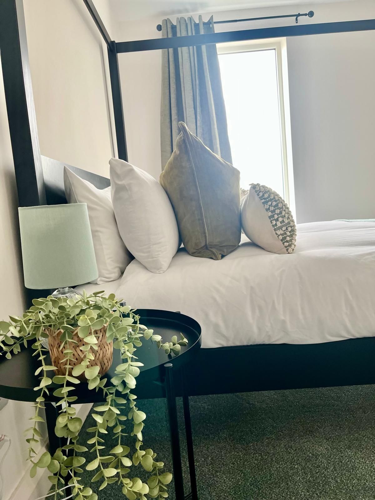 Neatly made bed with white pillows, gray accent pillow, and a bedside lamp beside a bright window.