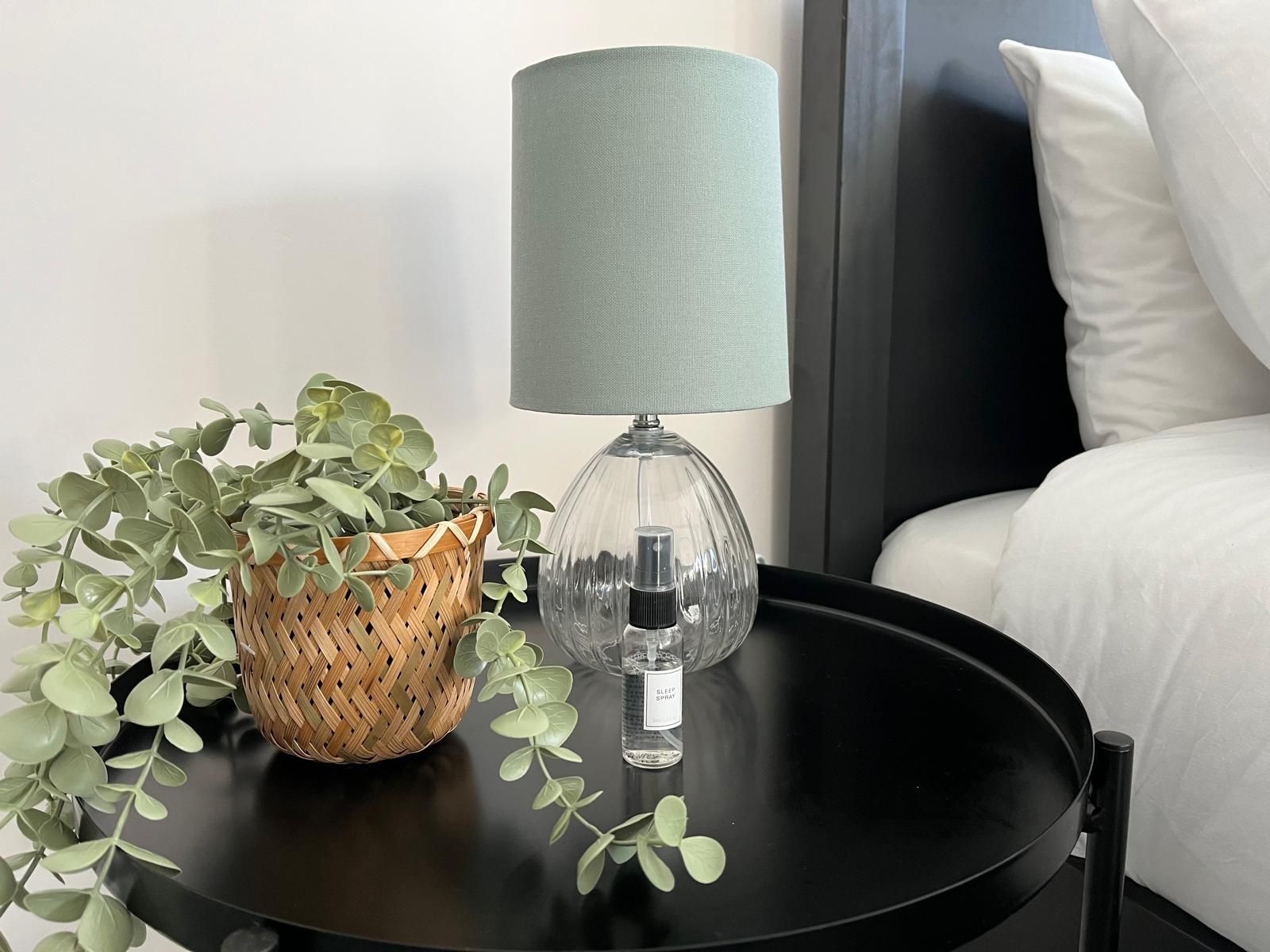 Black bedside table with a pale blue lamp and woven planter beside a white bed.