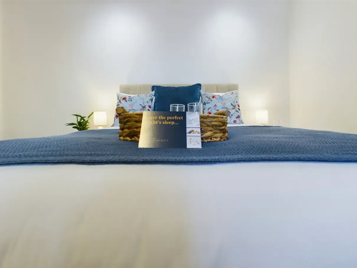 White bed with blue throw, patterned pillows, and warm bedside lamps in a minimalist bedroom