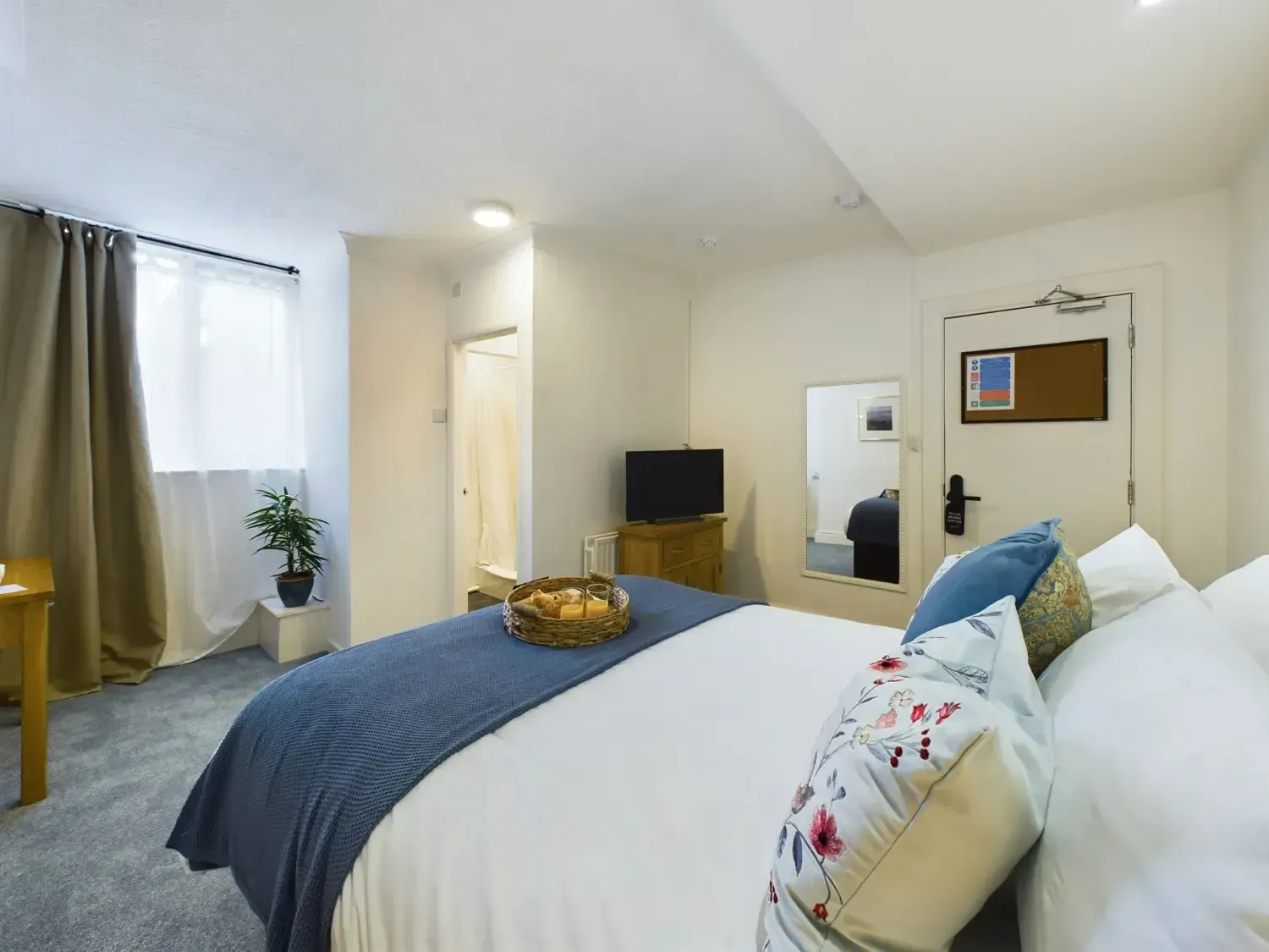Bright bedroom with a large bed, blue blanket, white walls, mirror, TV, and window curtains