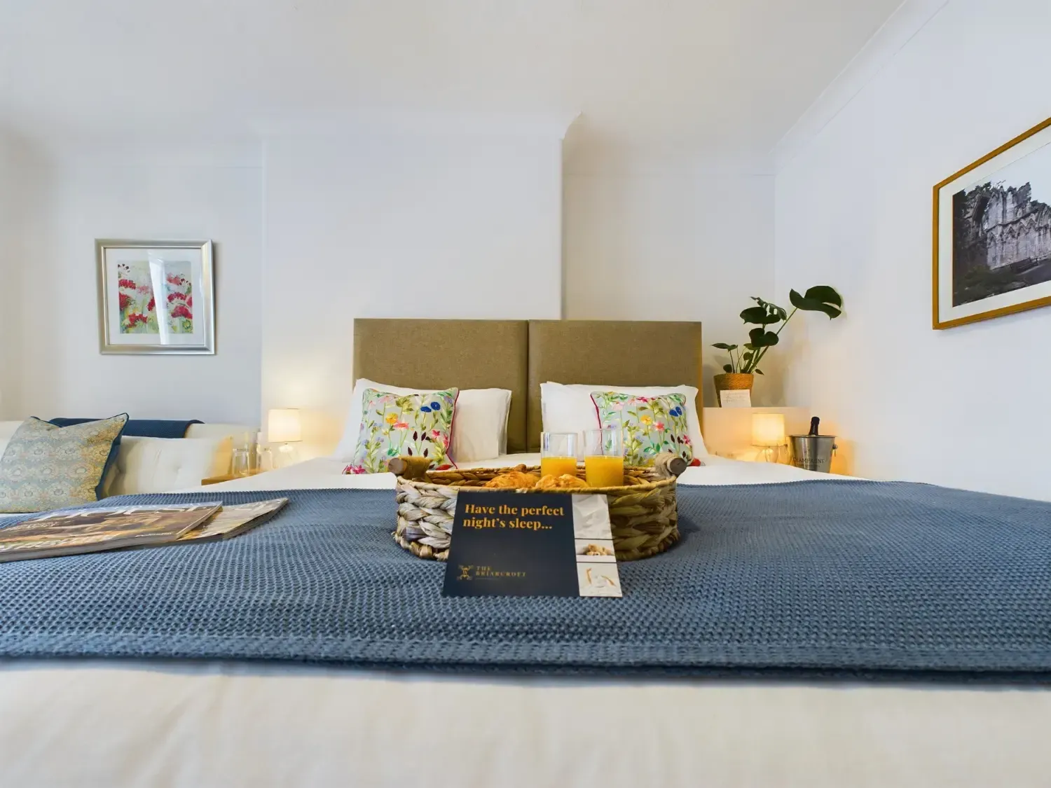 A neatly made bedroom with a bed, blue blanket, floral pillows, bedside lamps, and framed wall art.