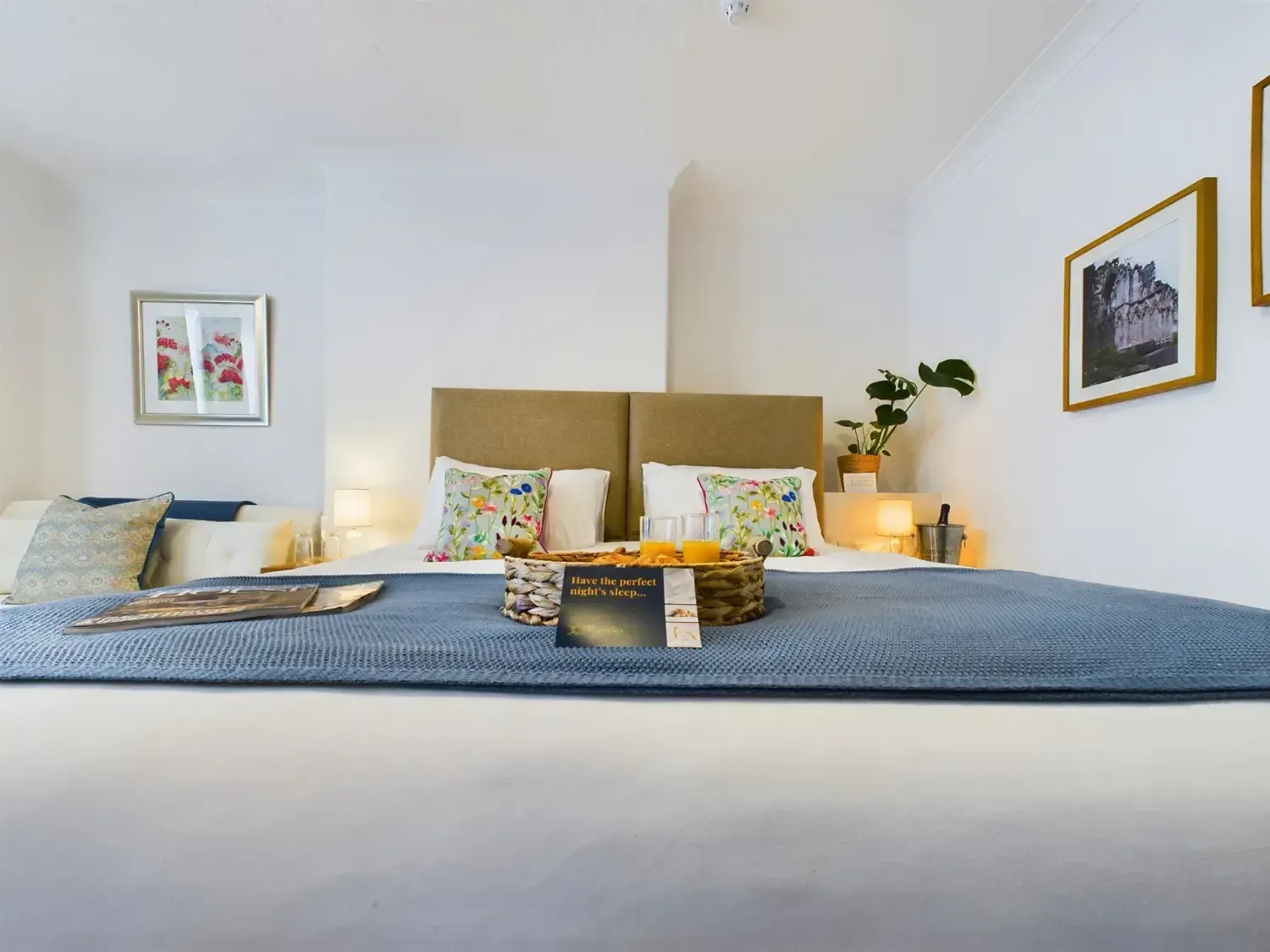 Cozy bedroom with a blue bedspread, patterned pillows, framed wall art, and warm bedside lighting