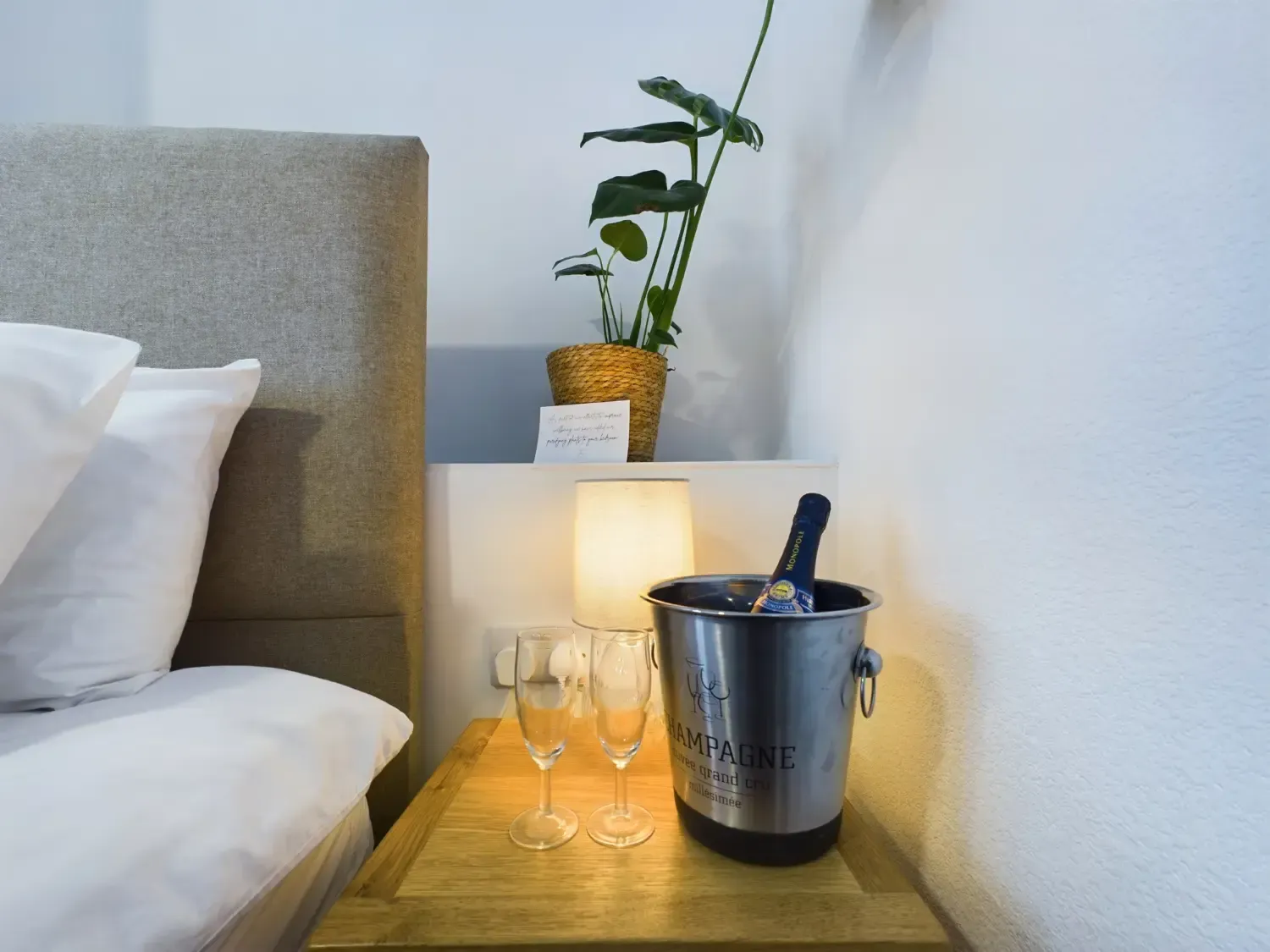 Bedroom nightstand with ice bucket, two wine glasses, lamp, and a potted plant beside the bed