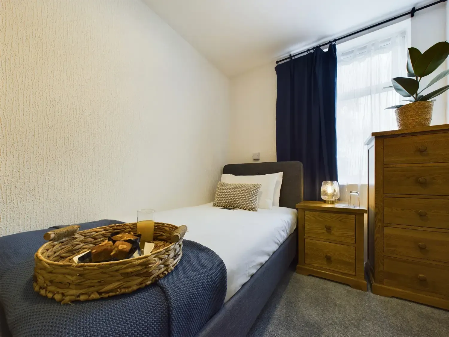 Cozy bedroom with a single bed, navy bedding, wicker tray, and wooden nightstand by a bright window.