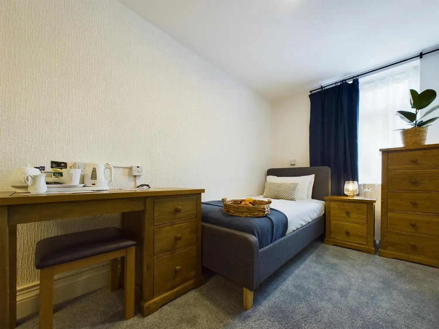 Small bedroom with a single bed, desk, blue curtains, and a dresser by a window.