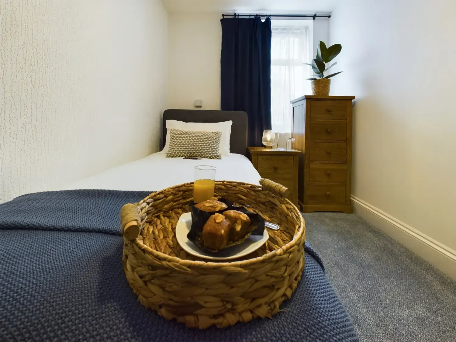 Small bedroom with a bed, wicker tray with breakfast, dresser, and window with dark curtains.