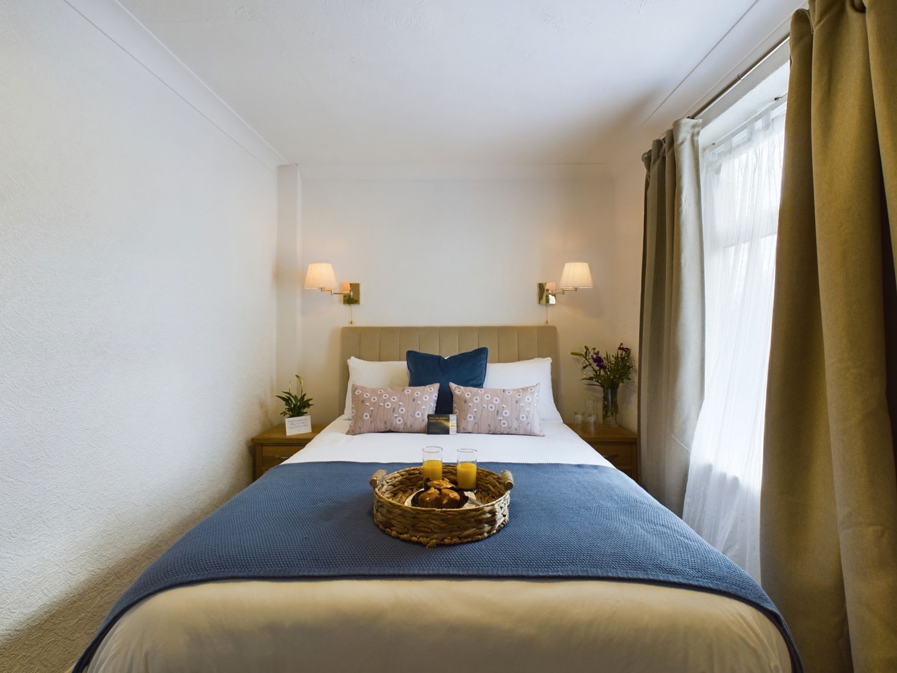 Cozy bedroom with a neatly made bed, blue throw, wall lamps, and a bright window with curtains.