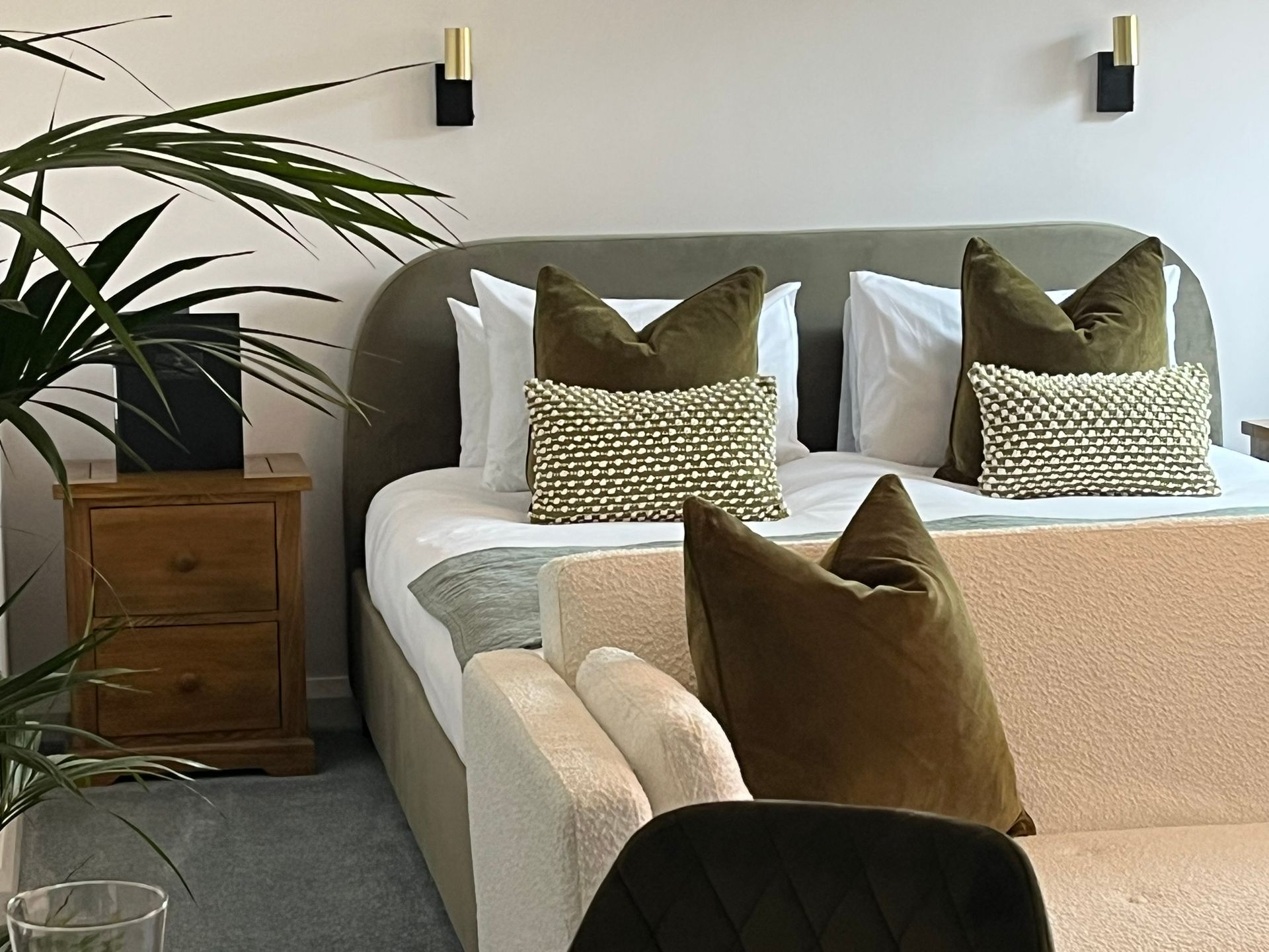 Bedroom with a neatly made bed, patterned pillows, wall sconces, and a wooden nightstand next to a potted plant.