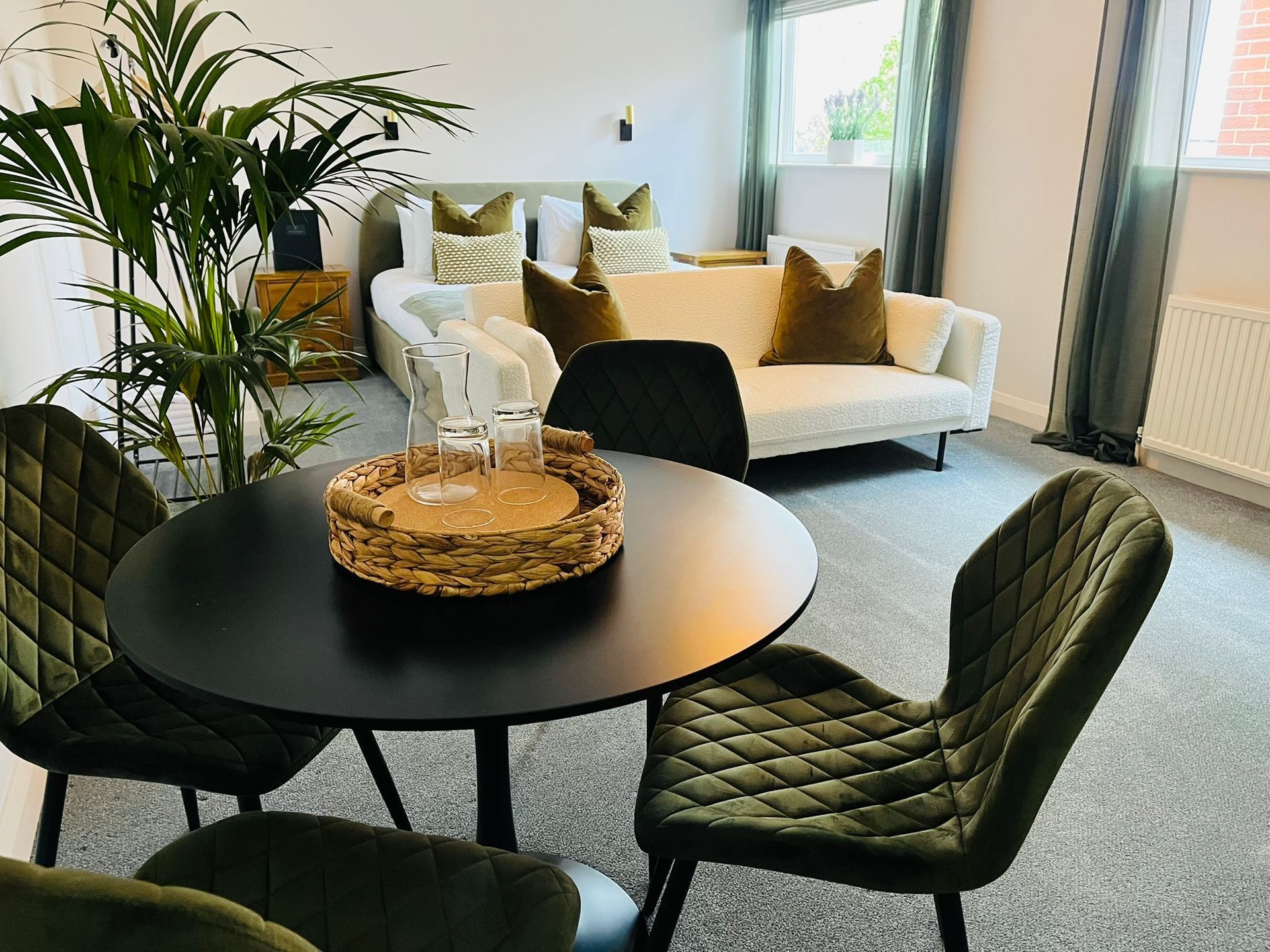 Modern living room with round black table, green chairs, white sofa, and large windows with plants.