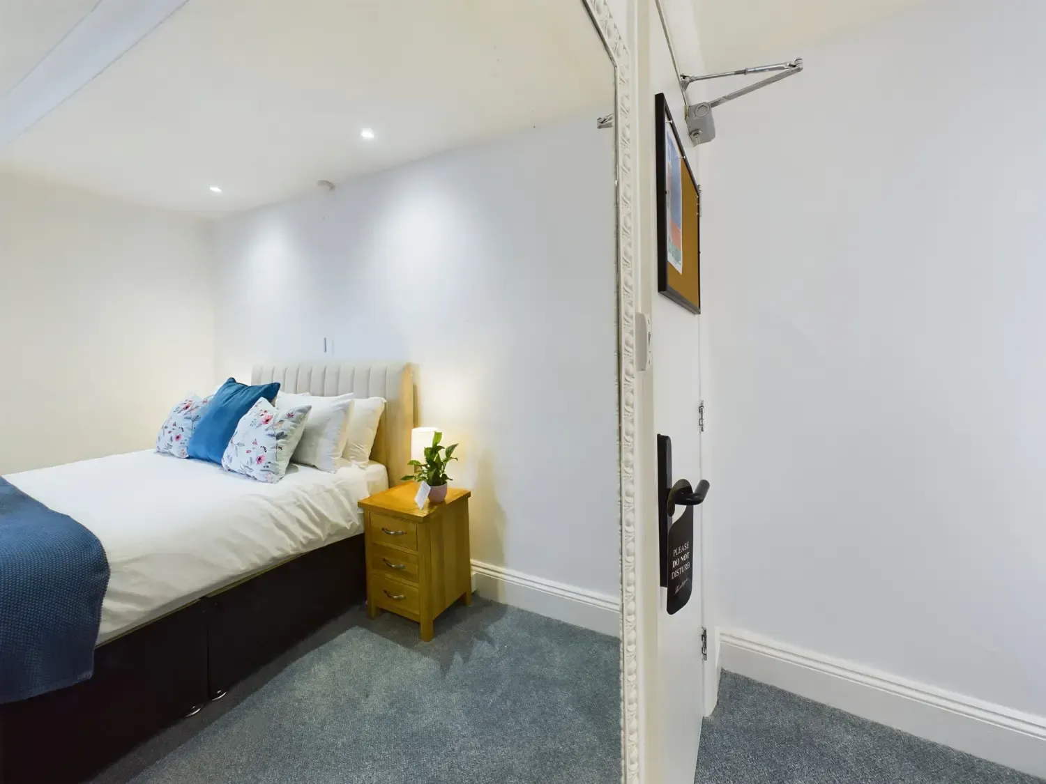 Small bedroom with a white bed, blue bedding, and a wooden nightstand beside a bright, open door.