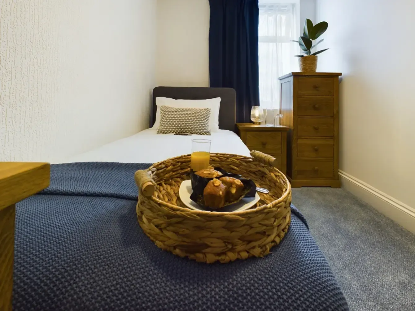 Cozy bedroom with a blue bedspread, woven basket tray, and wooden dresser by a window