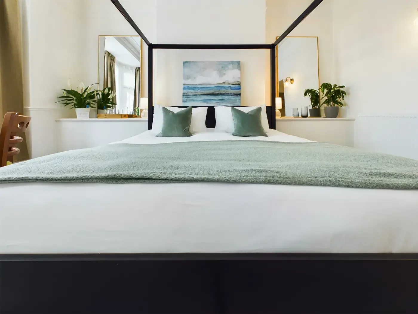 Bright bedroom with a white bed, green pillows, plants, and ocean artwork on the wall
