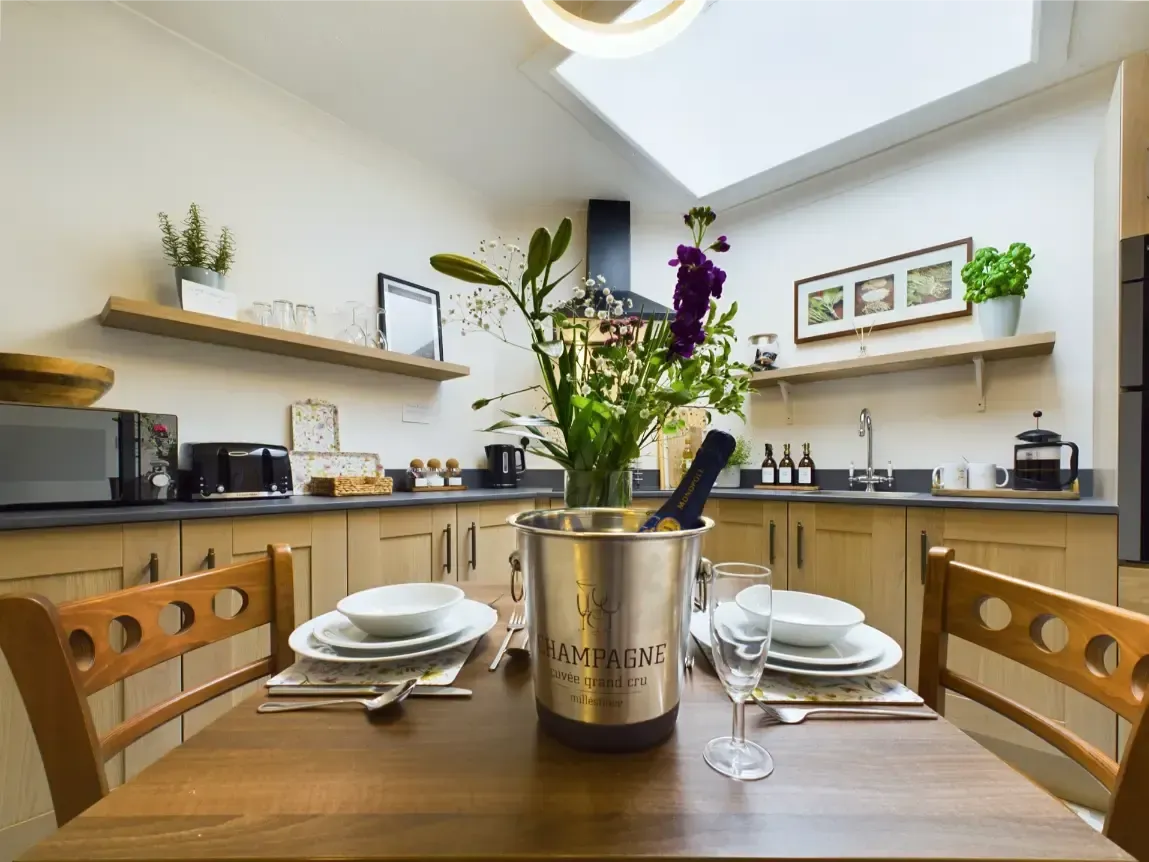 Modern kitchen with a set dining table, wooden chairs, and flowers in a metal vase.