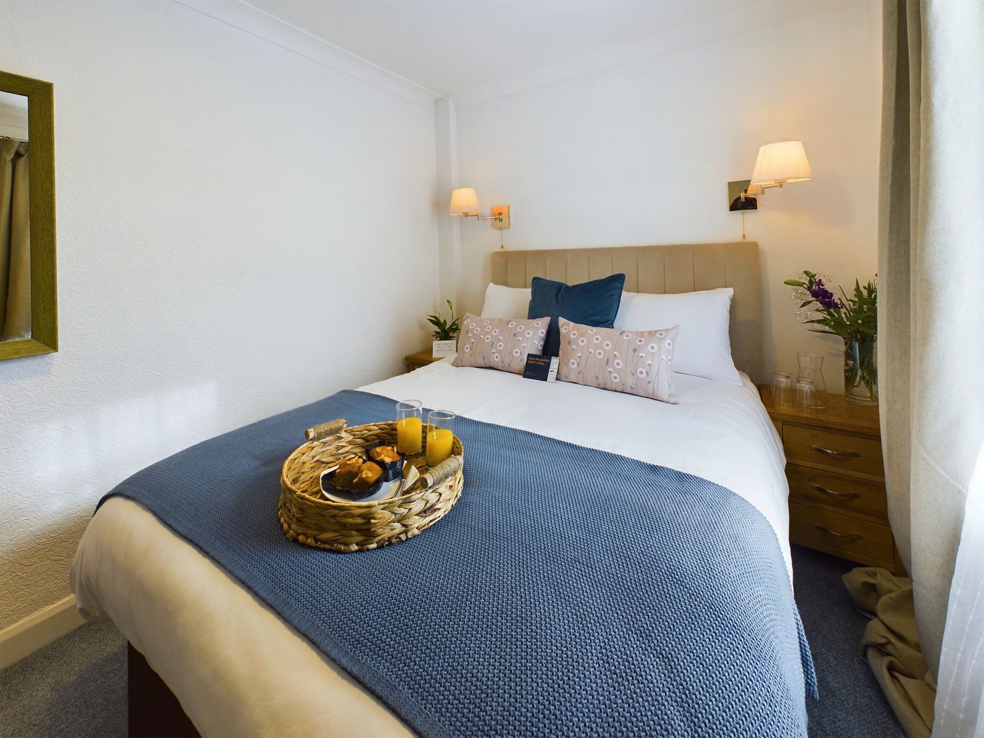 Cozy bedroom with a neatly made bed, blue quilt, bedside lamps, and a tray of fruit on the bed
