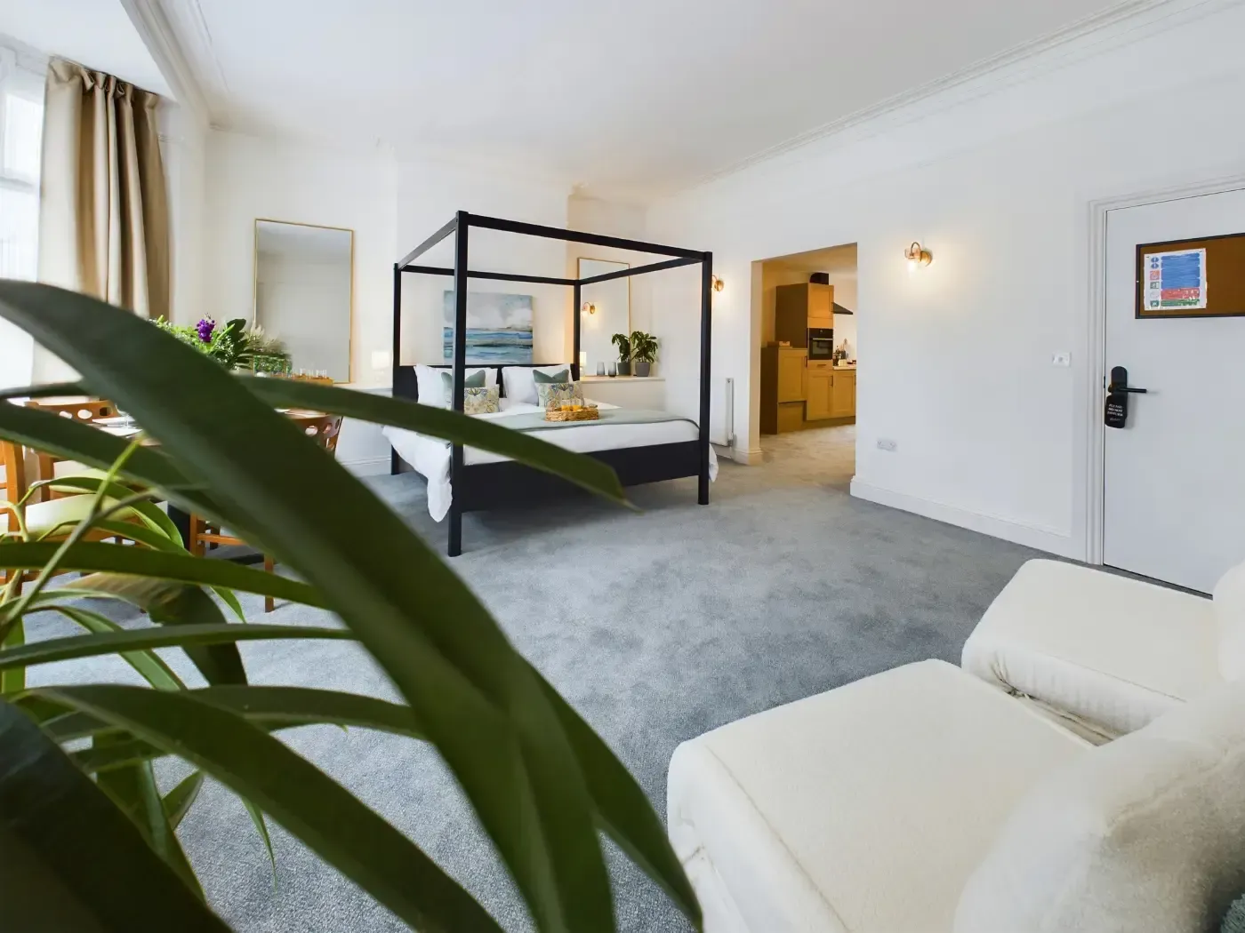 Bright bedroom with black canopy bed, white armchair, gray carpet, and plants near a window