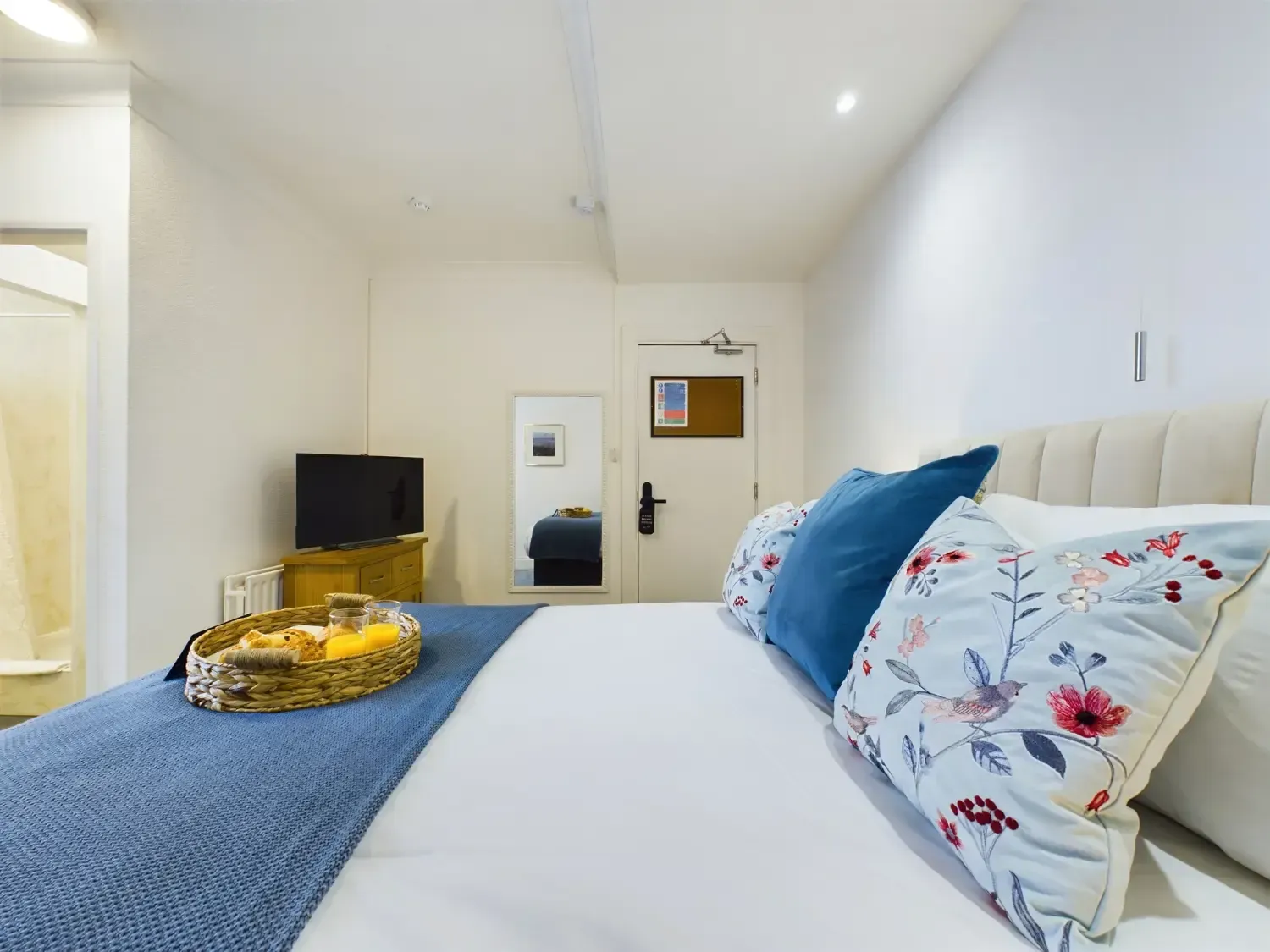 Bright bedroom with a white bed, blue blanket and floral pillows, TV, and a small golden tray on the bed