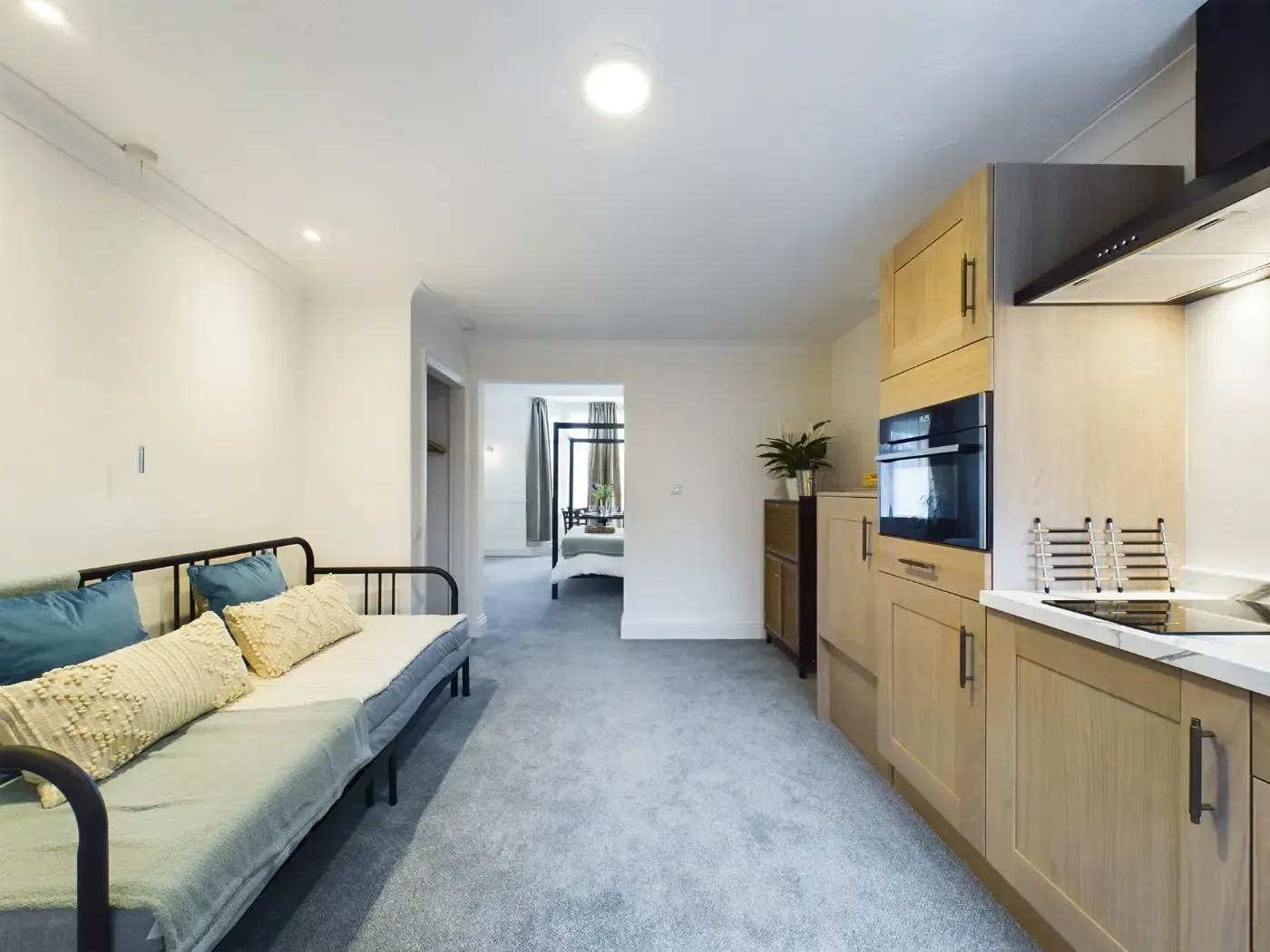 Bright studio room with gray sofa, kitchenette, TV, and hallway.