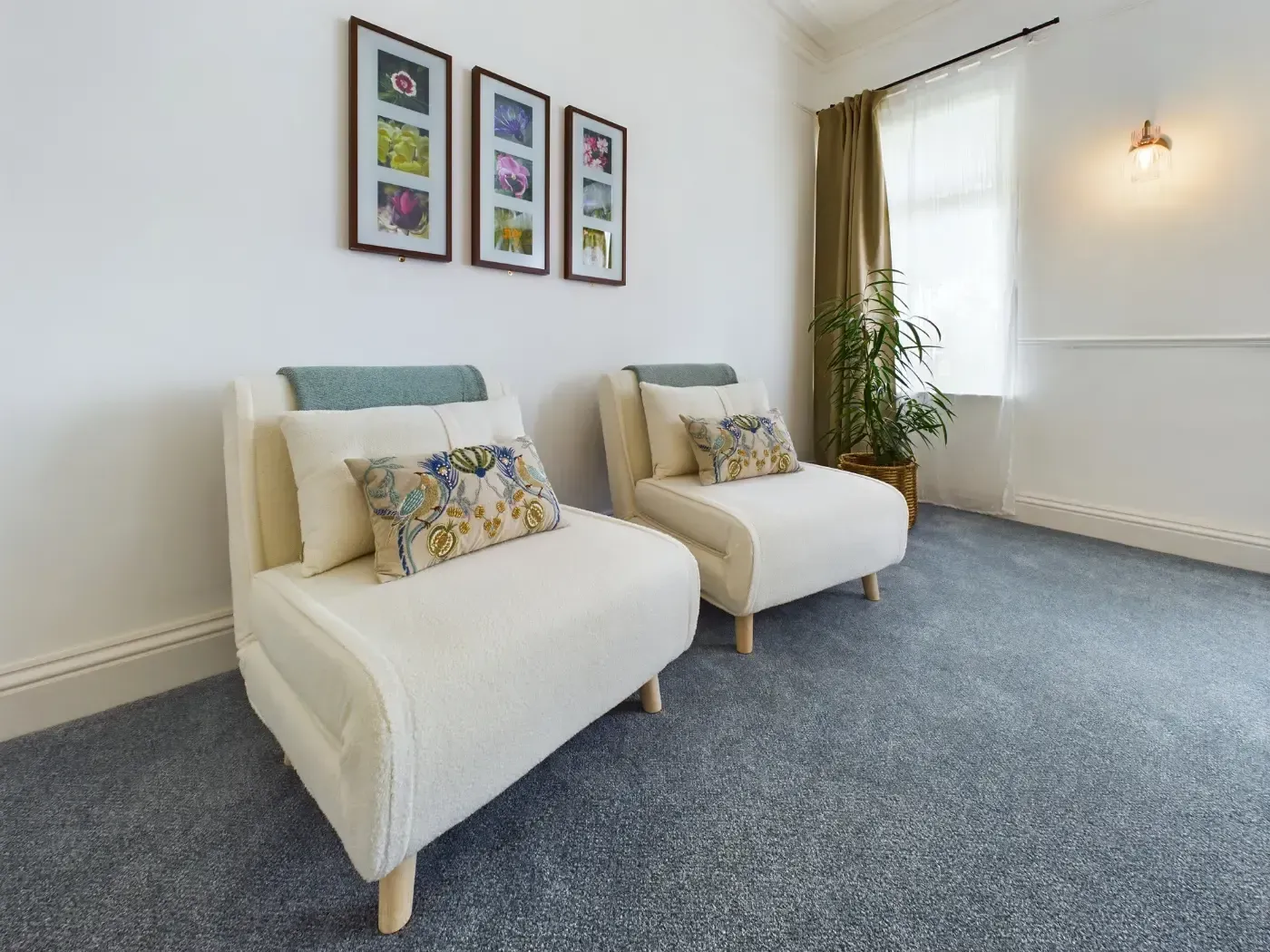 Bright living room with two cream chairs, framed art, blue carpet, and a window with curtains.