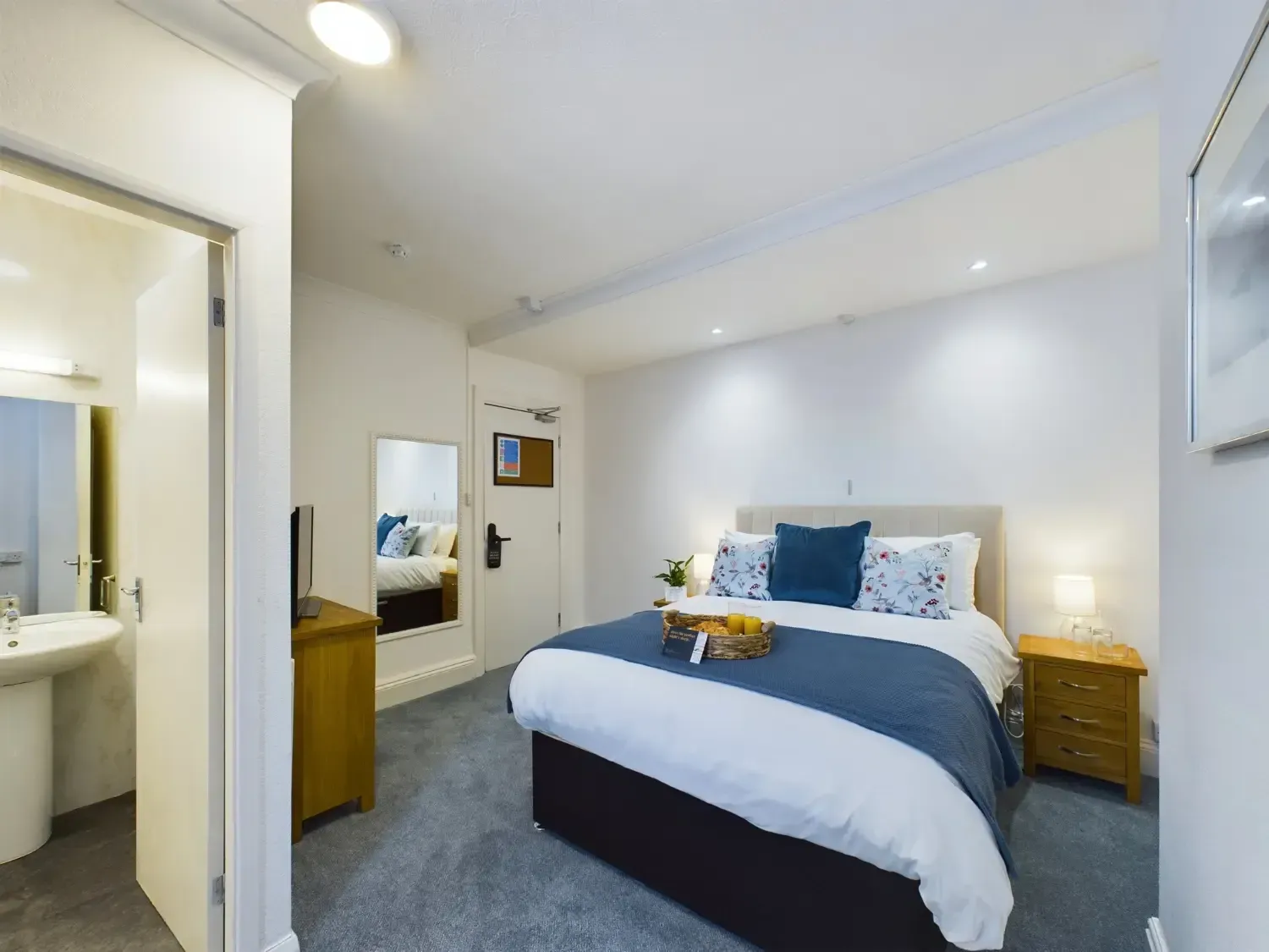 Bright modern bedroom with a neatly made bed, blue accents, nightstands, mirror, and an attached bathroom visible.