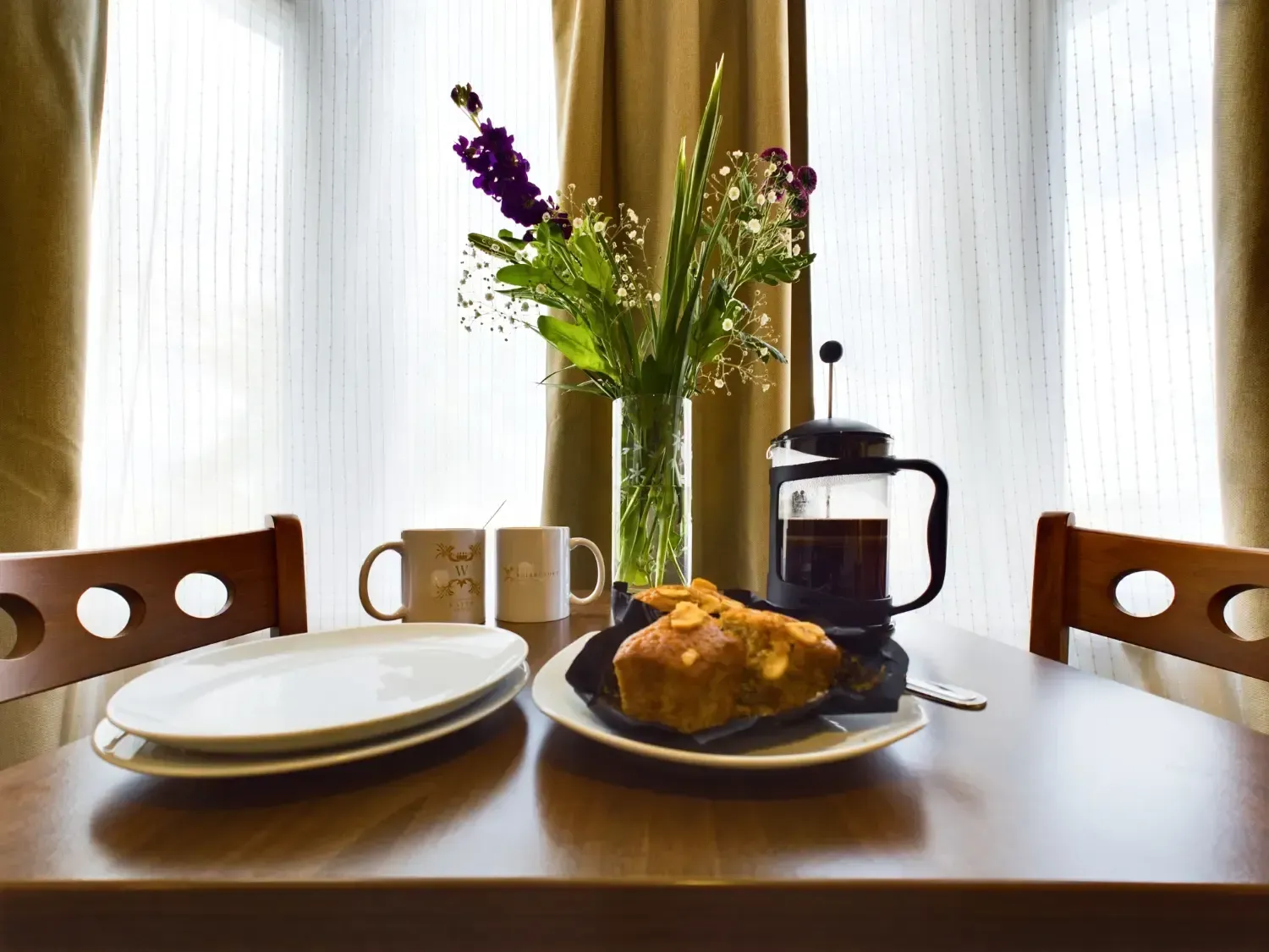Breakfast on a wooden table with plates, mugs, French press, and a vase of flowers by a bright window