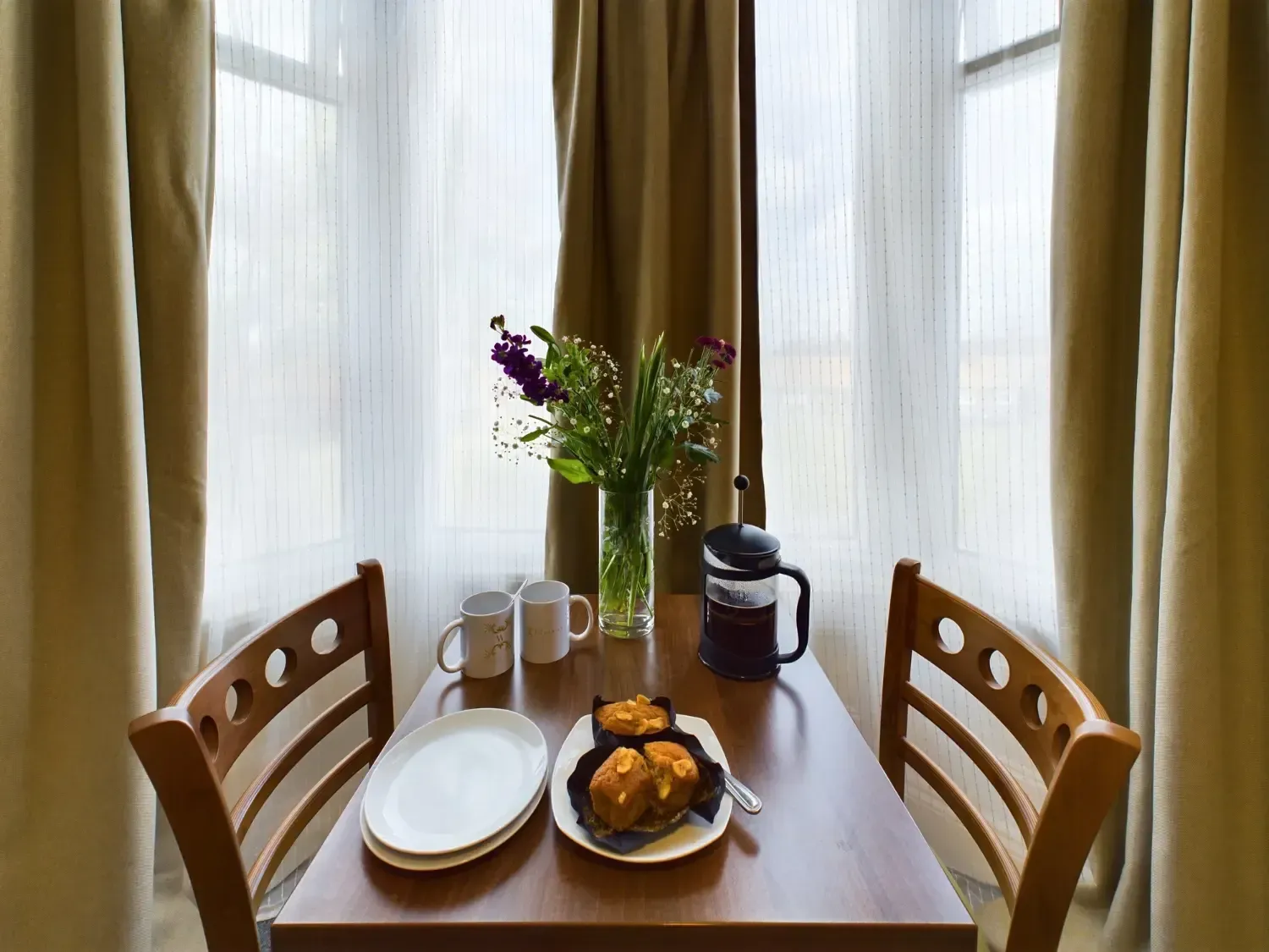 Small dining table by a window with curtains, plates, mugs, flowers, and a plate of fruit or pastries