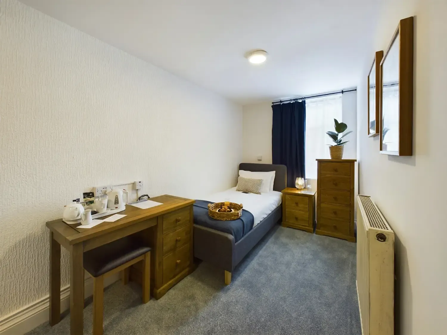Small bedroom with a single bed, desk, dresser, radiator, and blue curtains beside a window.