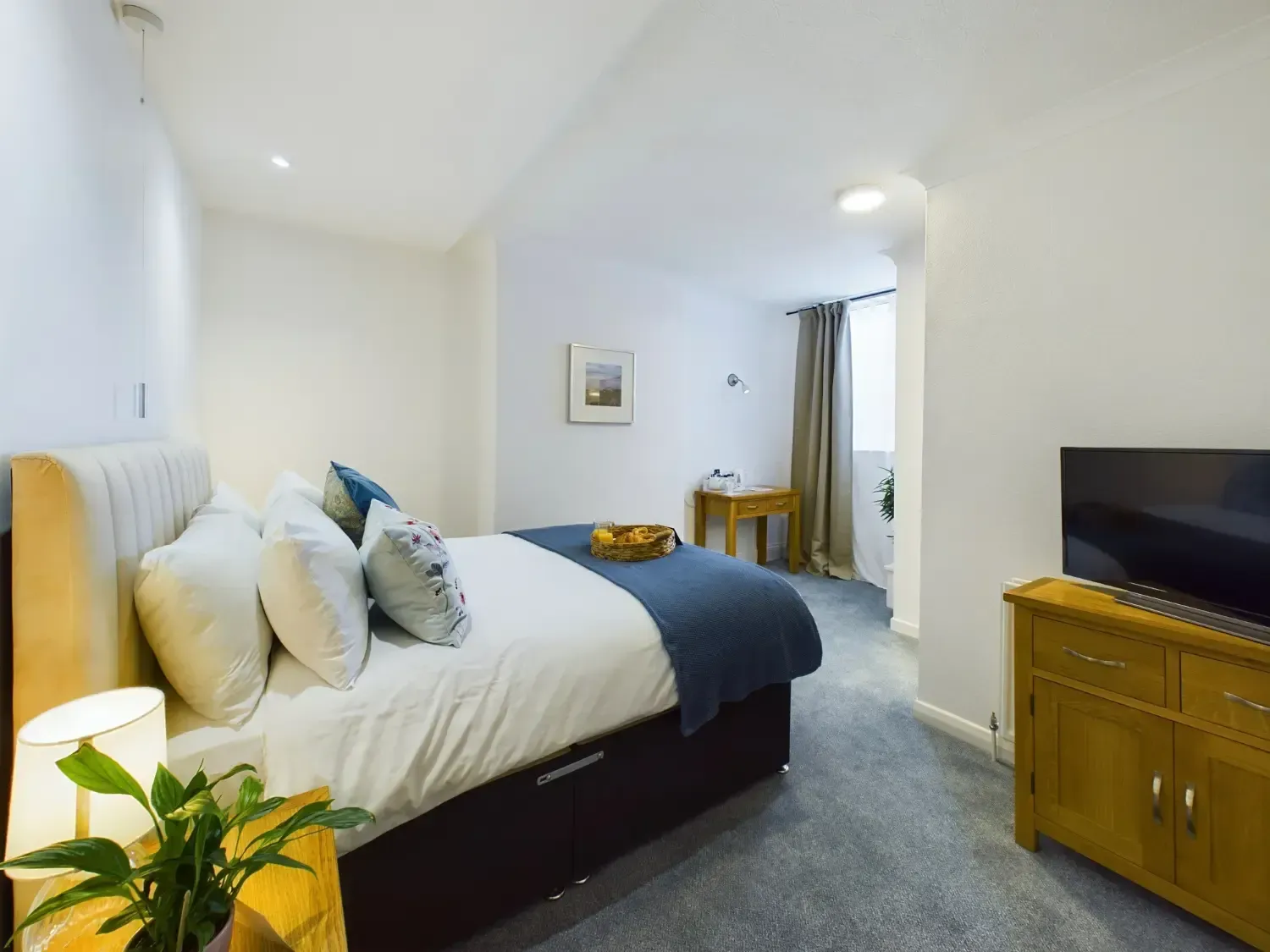 Bright bedroom with bed, TV stand, gray carpet, and a small desk near the window