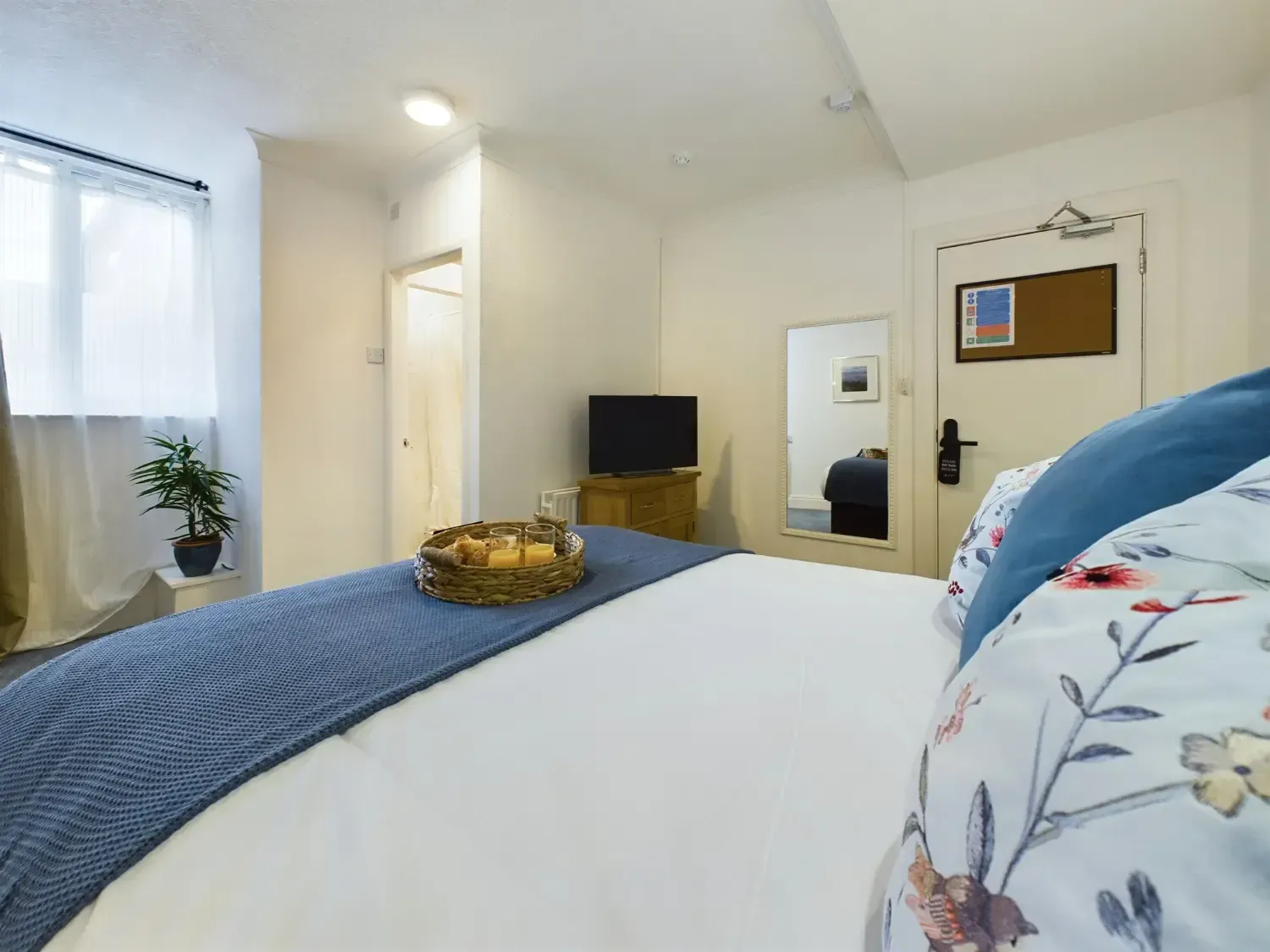 Bright bedroom with white walls, blue bedding, window light, TV, mirror, and a small table with decor.