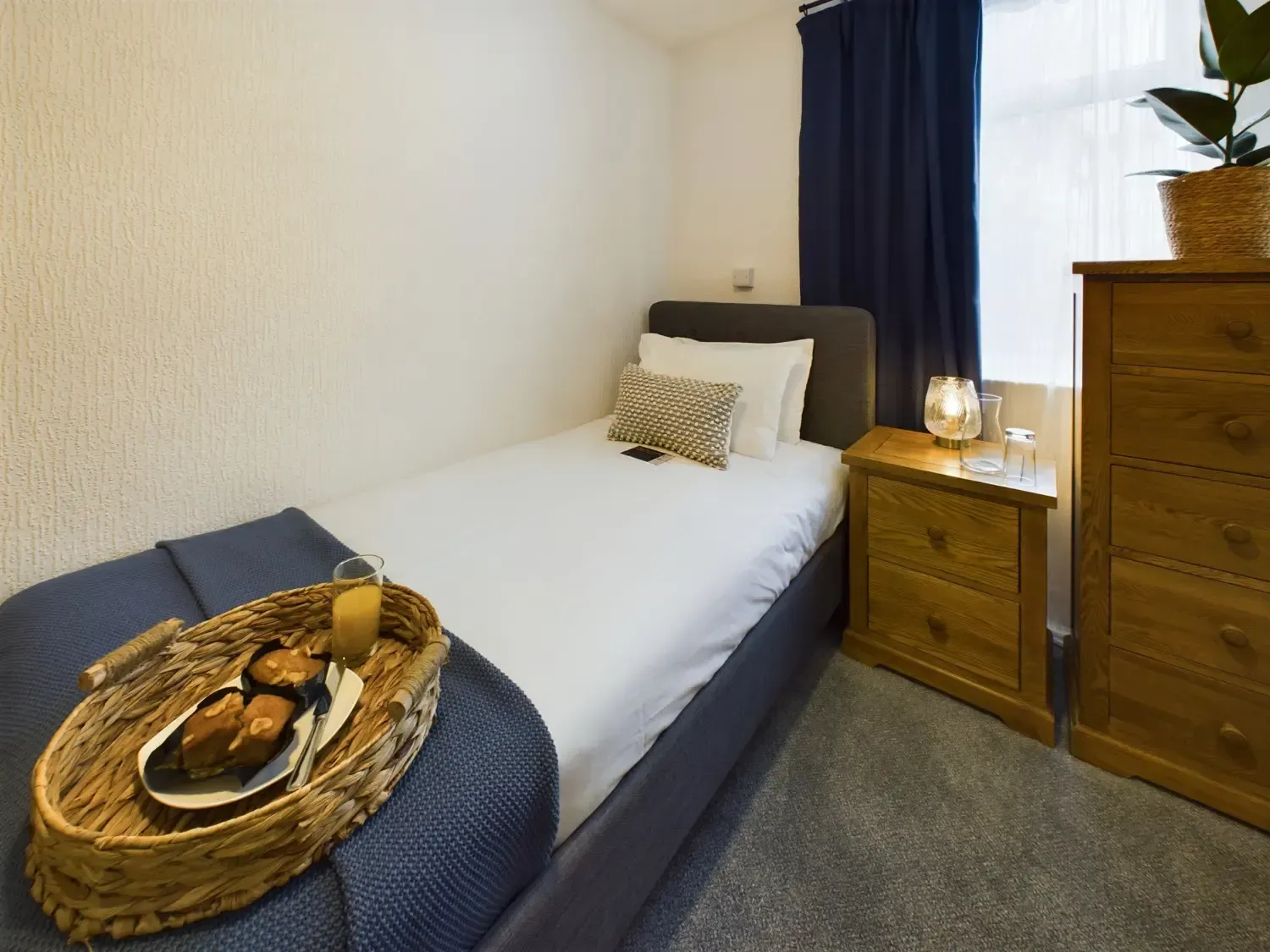 Small bedroom with a single bed, blue bedding, wooden nightstand, and dresser by a window with dark curtains.