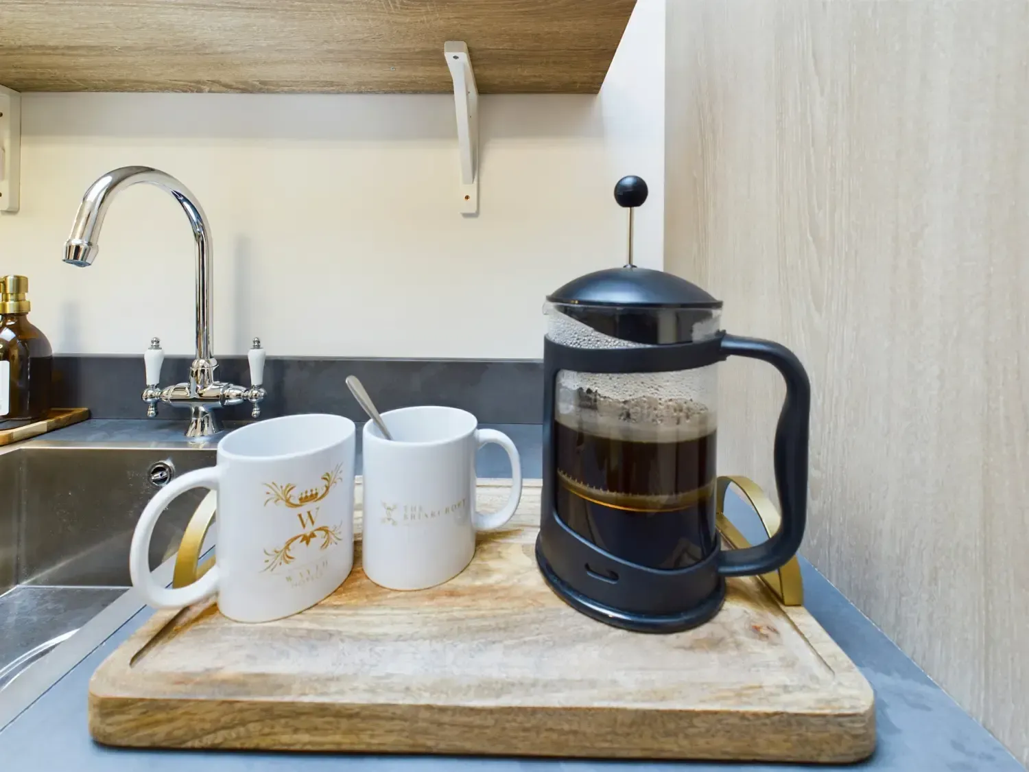 French press coffee and two mugs on a wooden tray beside a kitchen sink