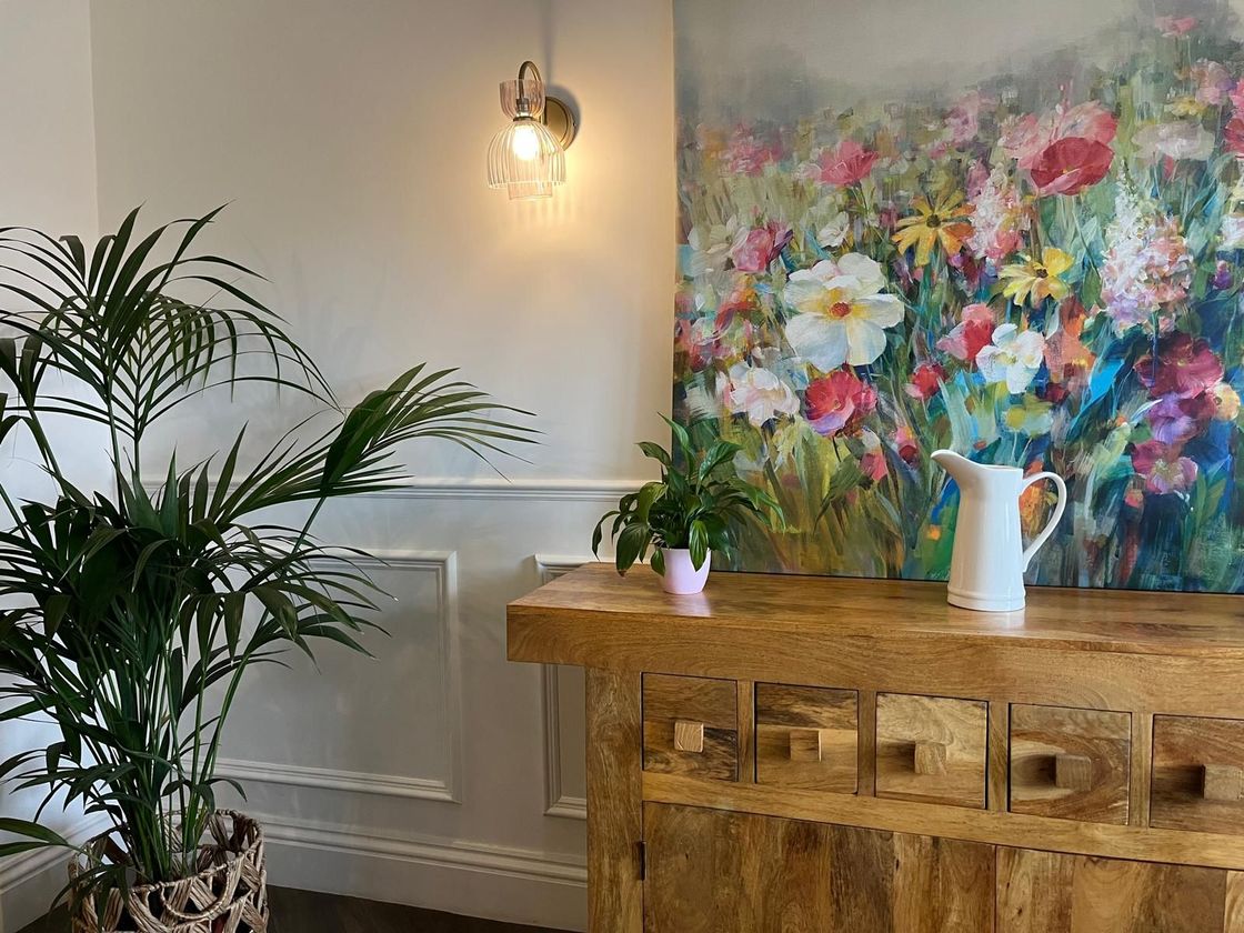 Bright room with a large colorful floral painting, wooden cabinet, plant, and wall sconce.
