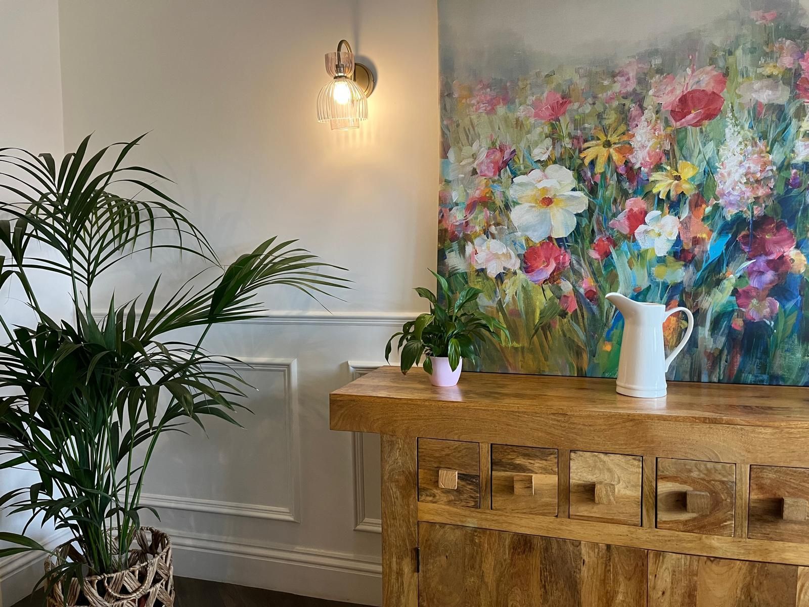 Bright room with a large colorful floral painting, wooden cabinet, plant, and wall sconce.