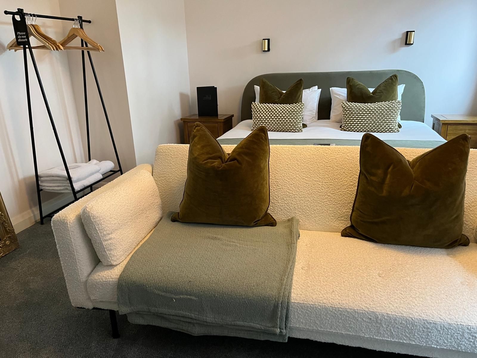 Cozy bedroom with a bed, patterned pillows, olive sofa, and black floor lamp in soft neutral tones